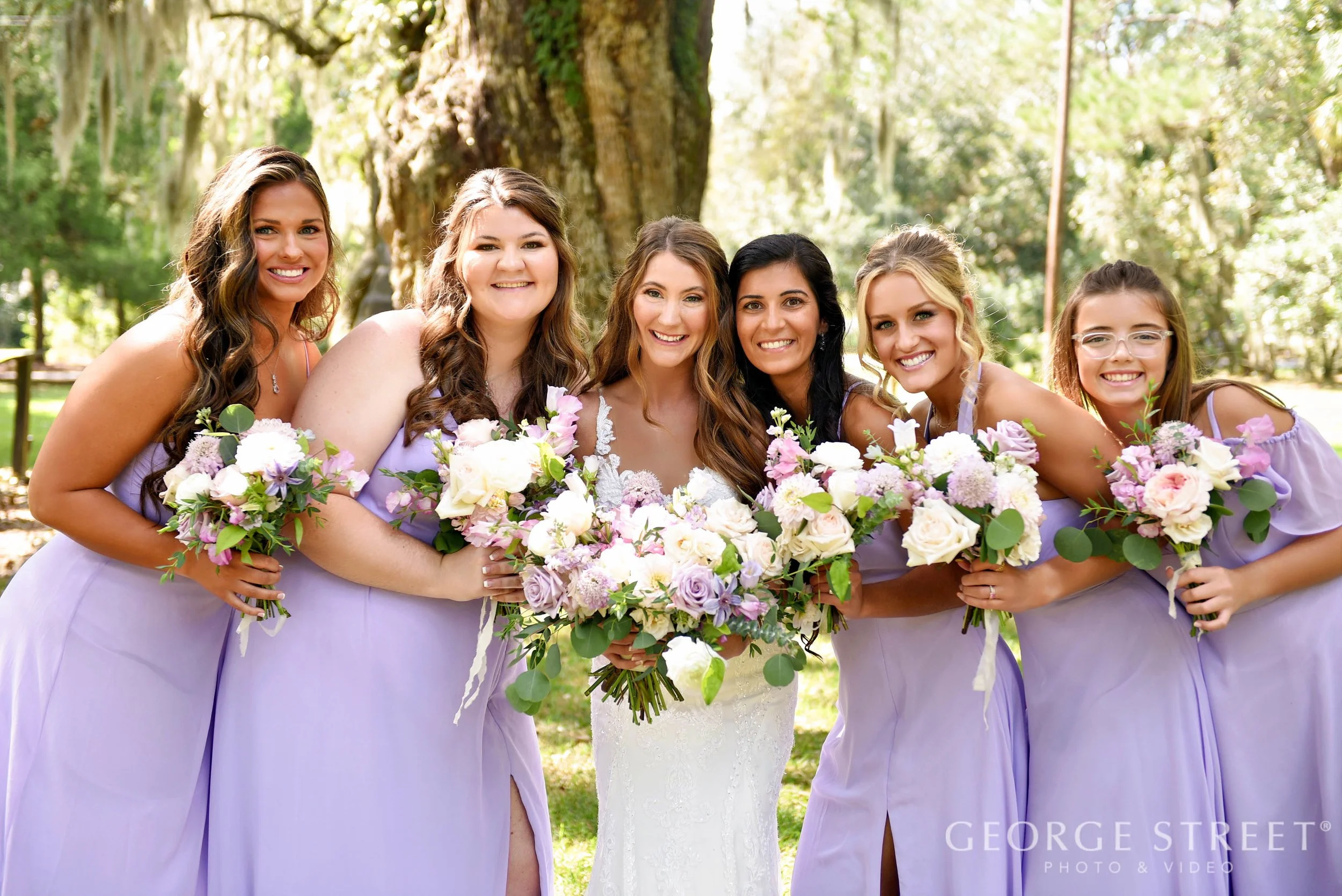 Six women in lavender bridesmaid dresses and a bride in white wedding gown holding bouquets of pink, white, and purple flowers, smiling outdoors under a large tree in a park.
