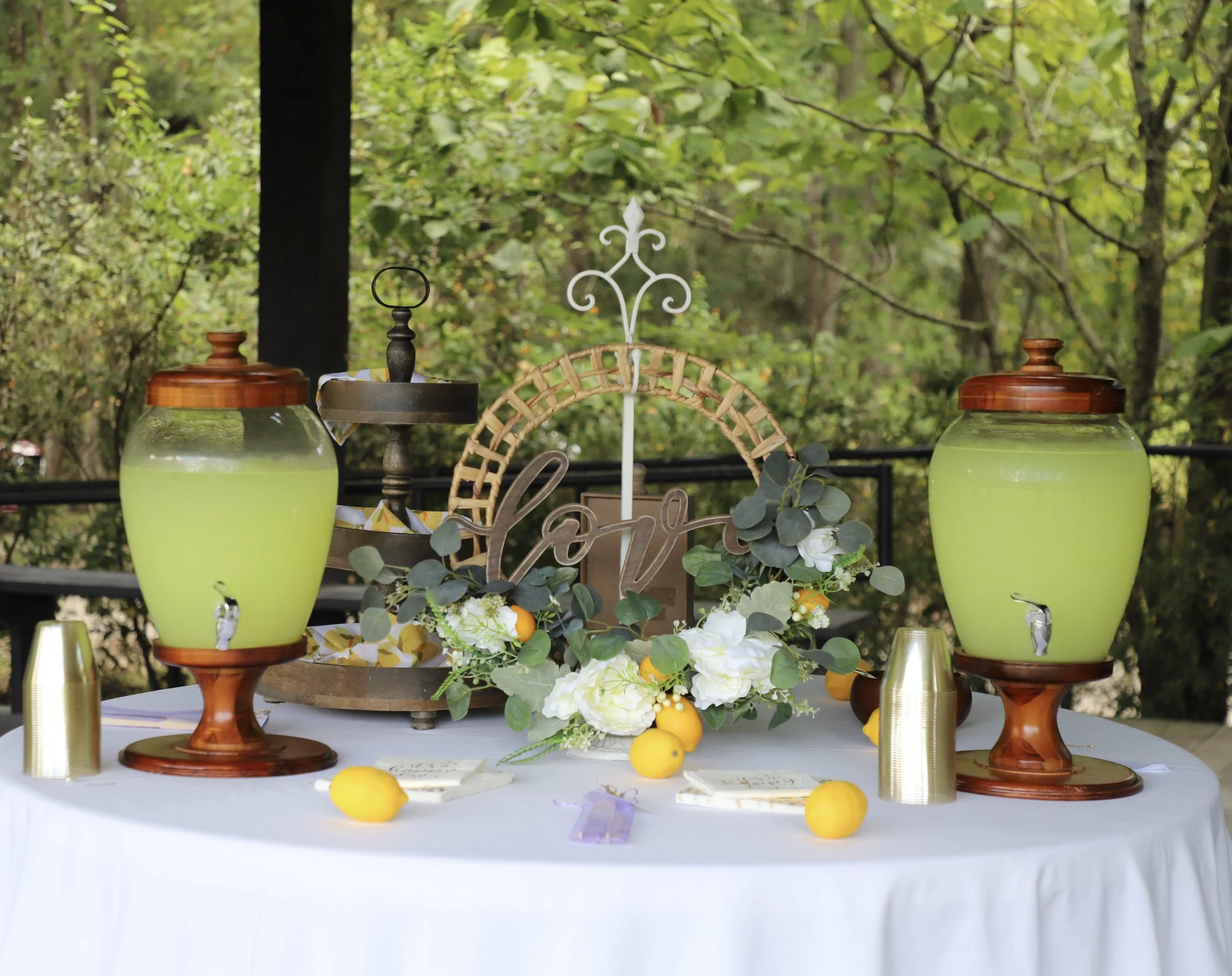 Table set outdoors with two large glass beverage dispensers filled with yellow-green drinks, surrounded by lemons, white flowers, and greenery, with a decorative lace and metal centerpiece and plates.