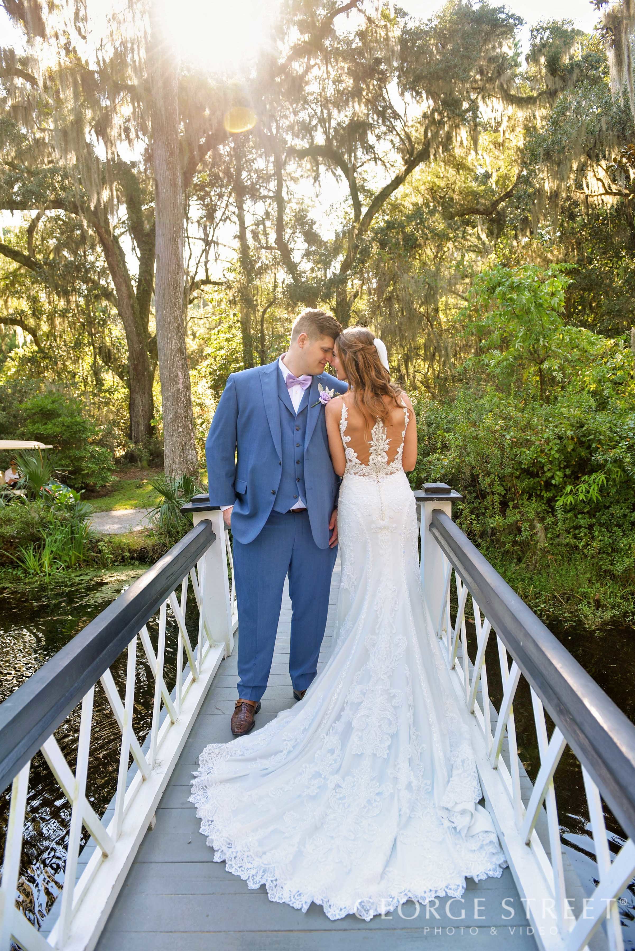 A newly married couple stands on a small white bridge in a lush green outdoor setting during sunset, sharing an intimate moment with foreheads touching.