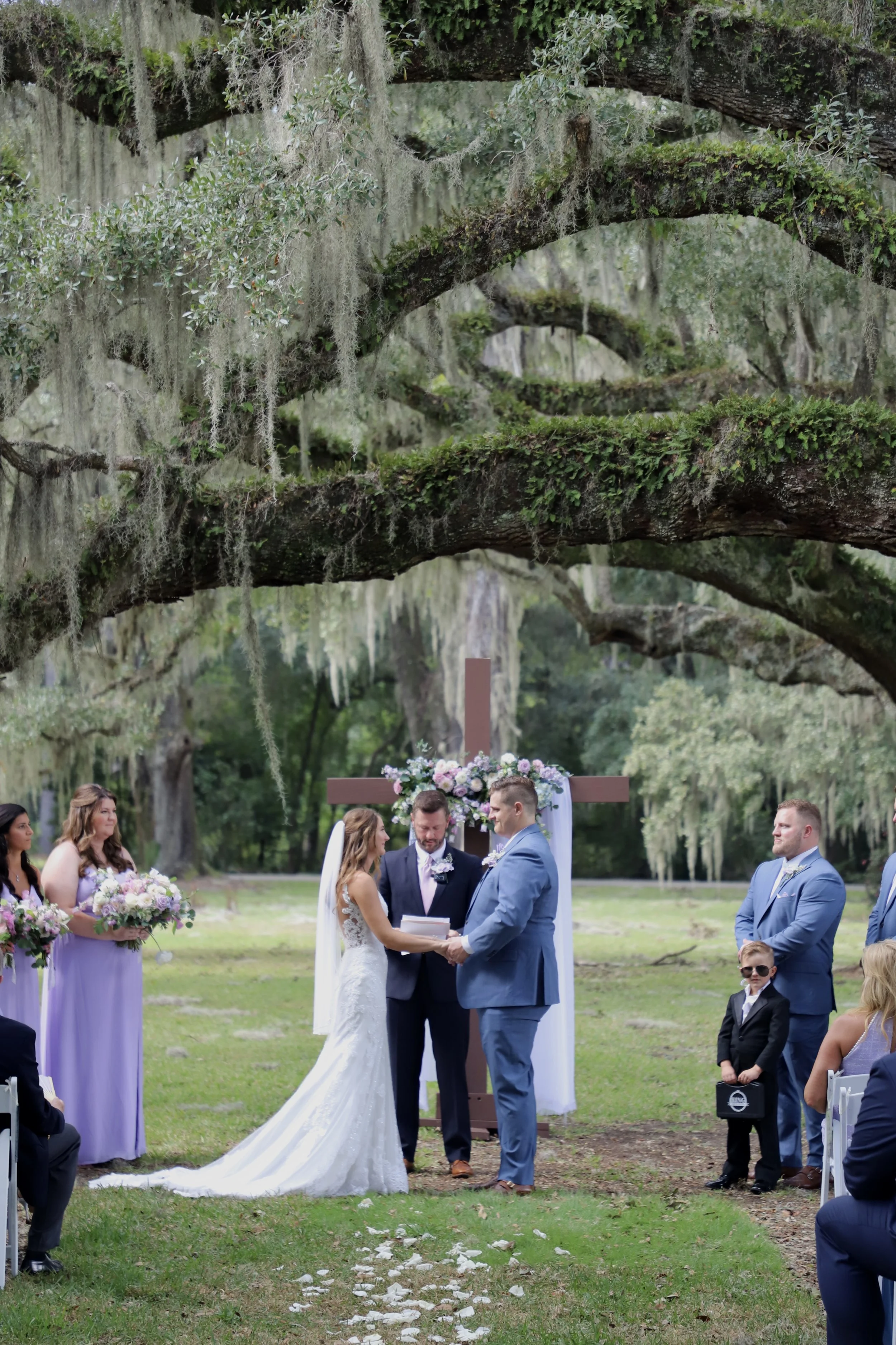 A wedding ceremony outdoors under large, moss-covered trees with hanging Spanish moss, with a bride and groom exchanging vows in front of an officiant and wedding party.