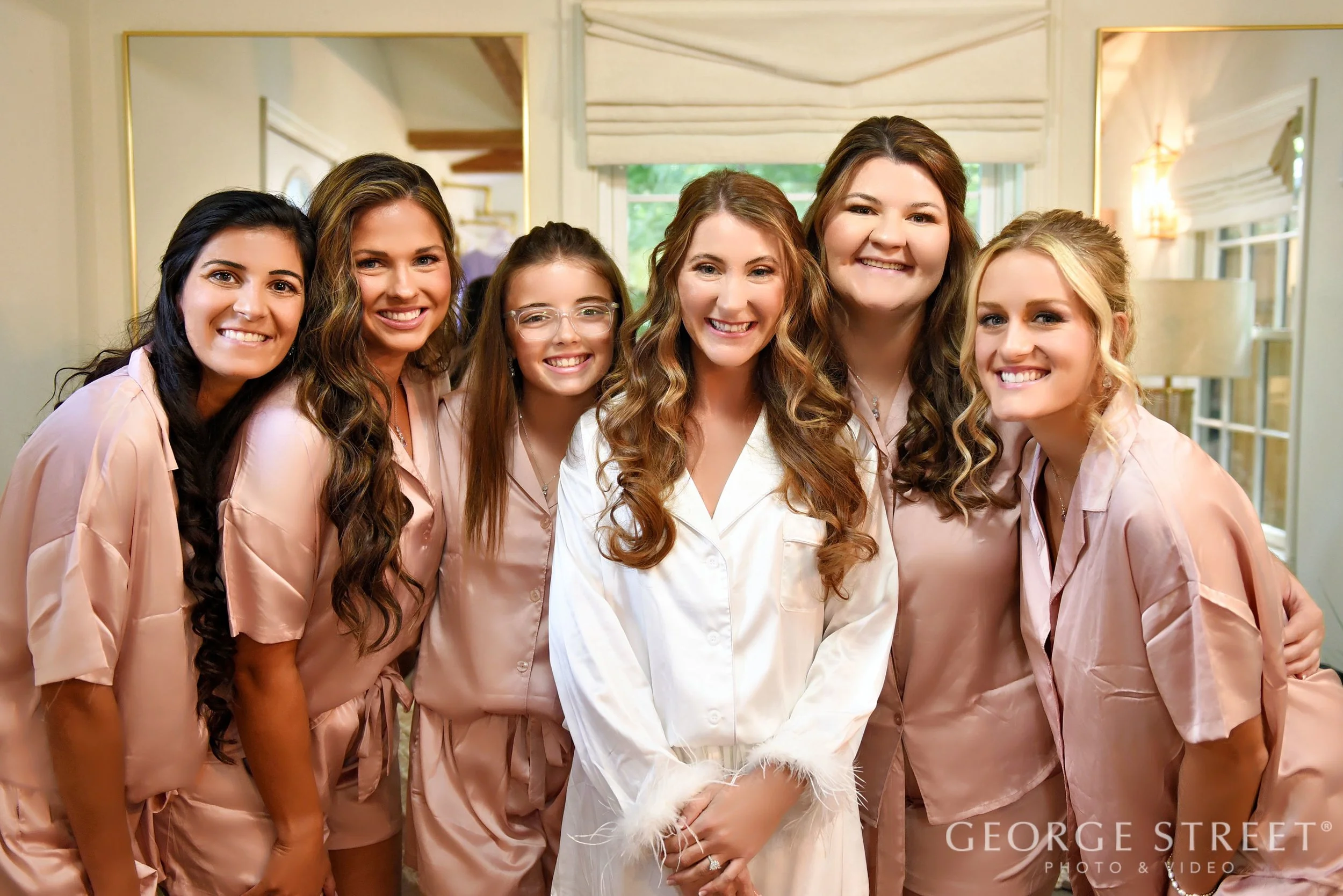 A group of seven women, dressed in satin pajamas, smiling and posing together indoors with bright natural light, in a cozy and stylish room.