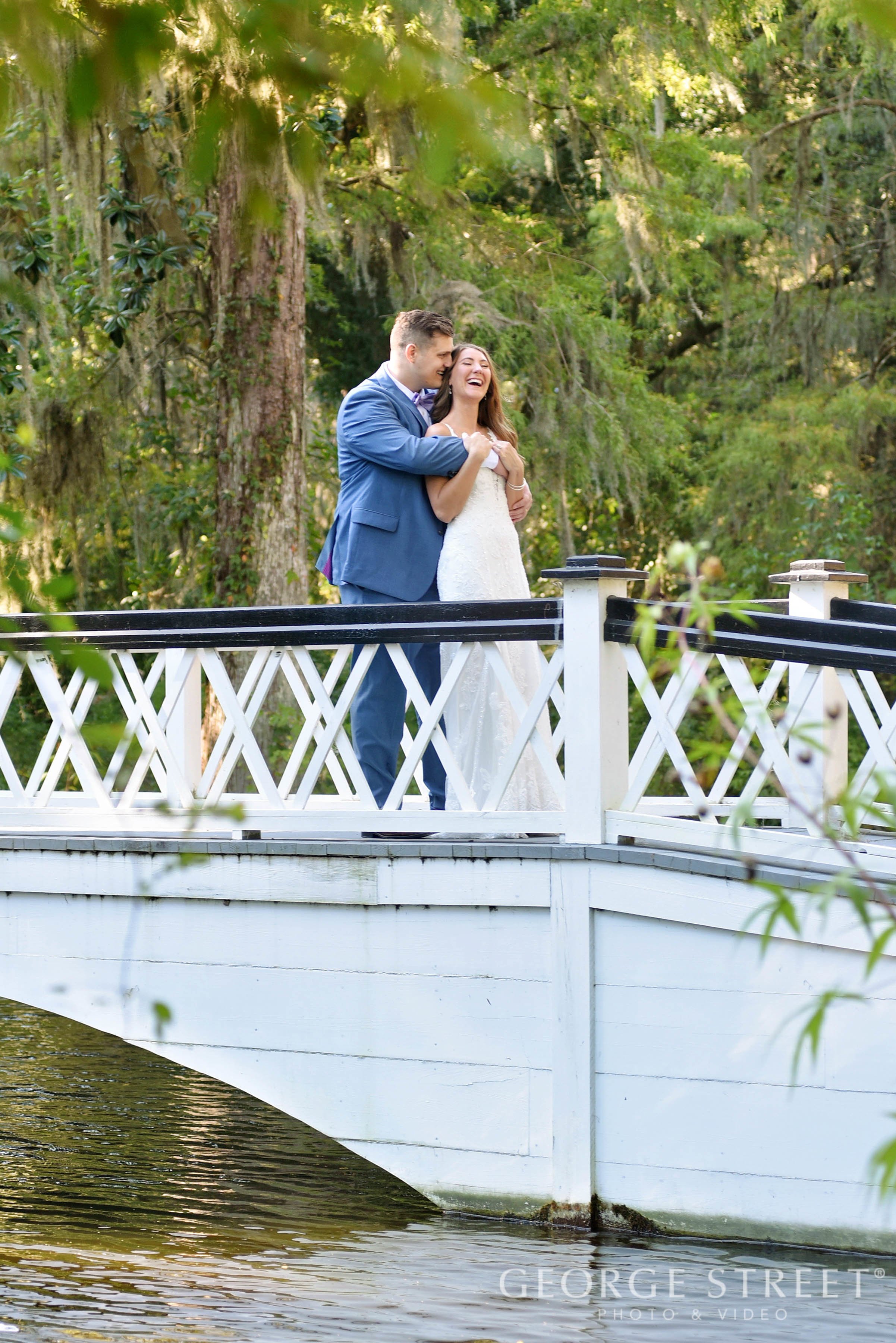 A bride and groom stand on a white bridge over water, embracing and laughing surrounded by lush green trees in a scenic outdoor setting.
