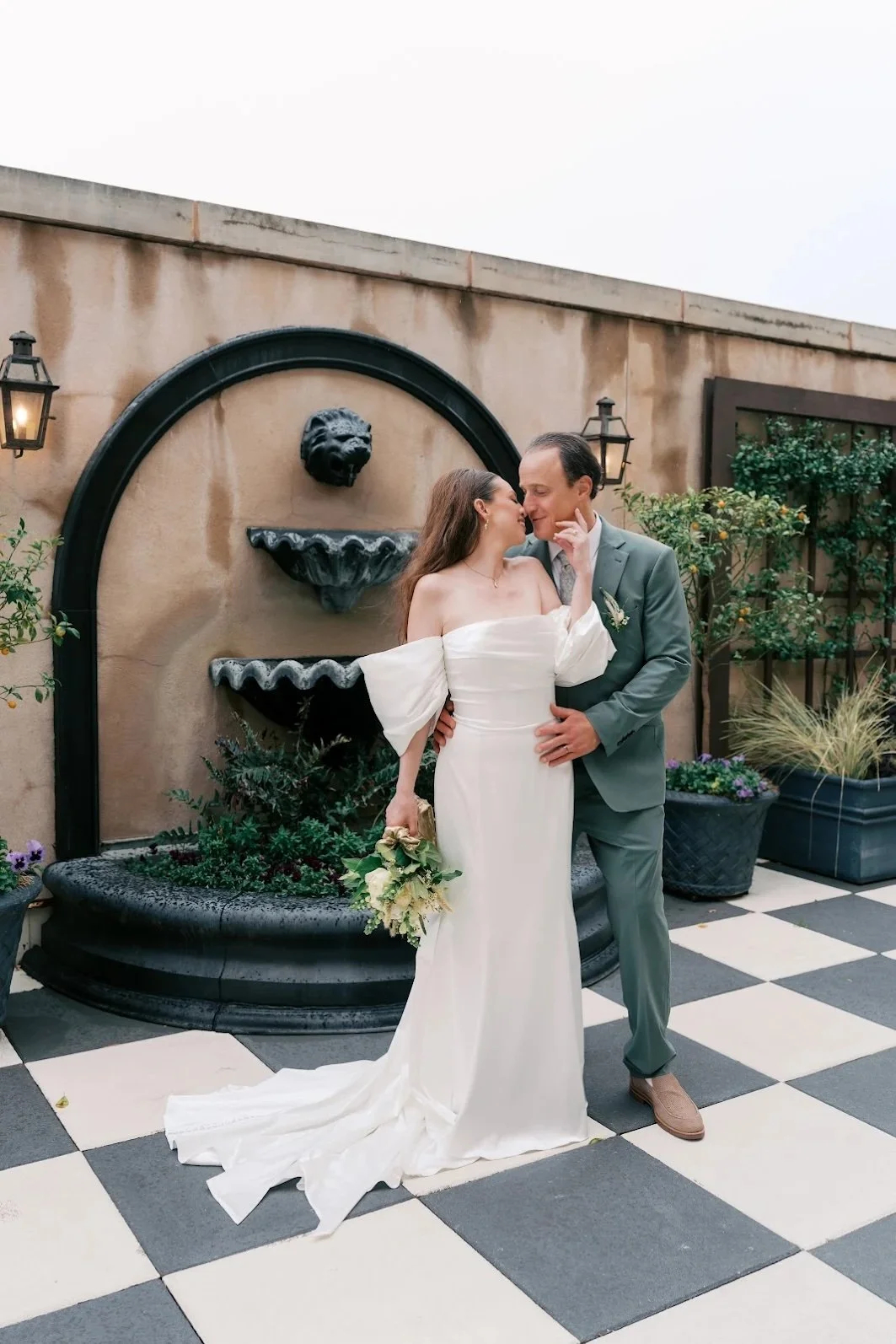 A bride and groom embrace and smile at each other on a checkered floor, with a fountain and potted plants in the background.