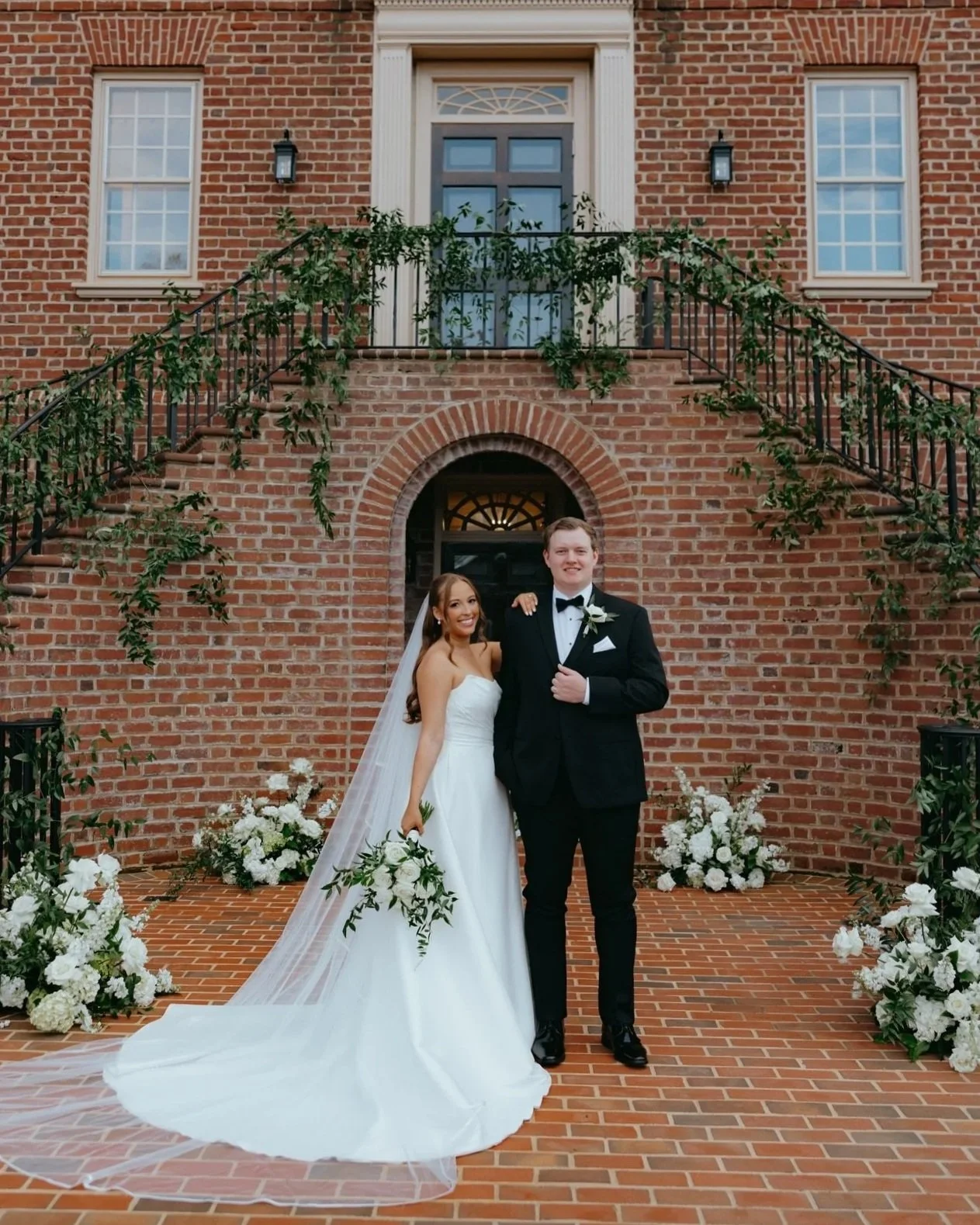 A bride and groom standing outside a brick building, smiling. The bride is in a white wedding gown holding a bouquet, and the groom in a black tuxedo with a bow tie. Flower arrangements are on either side of them.