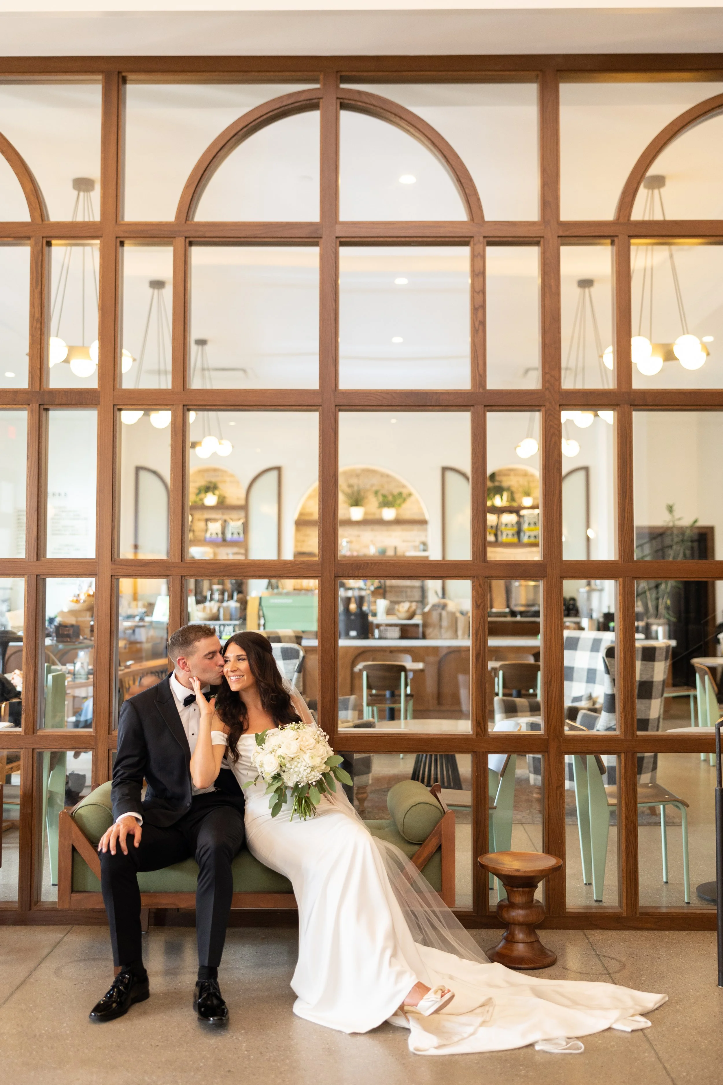 A newlywed couple sitting on a bench, with the groom kissing the bride's cheek as she holds a bouquet of white flowers in front of a decorative wooden partition in a restaurant or cafe setting.