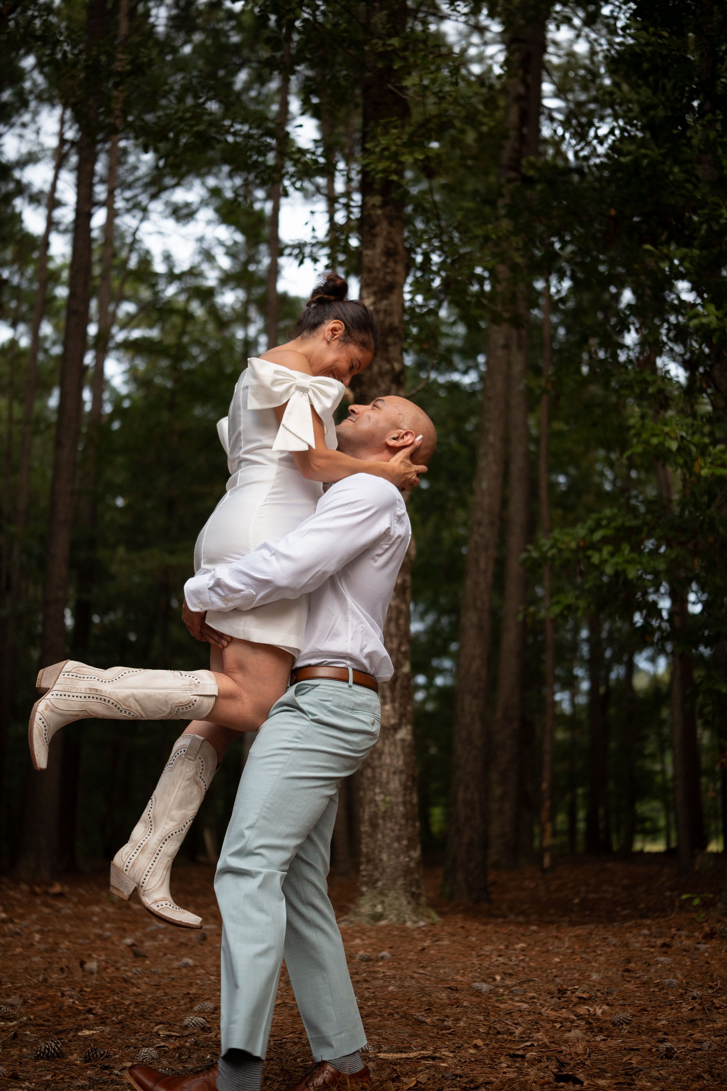Man lifting woman, both smiling, in a forest with tall trees.