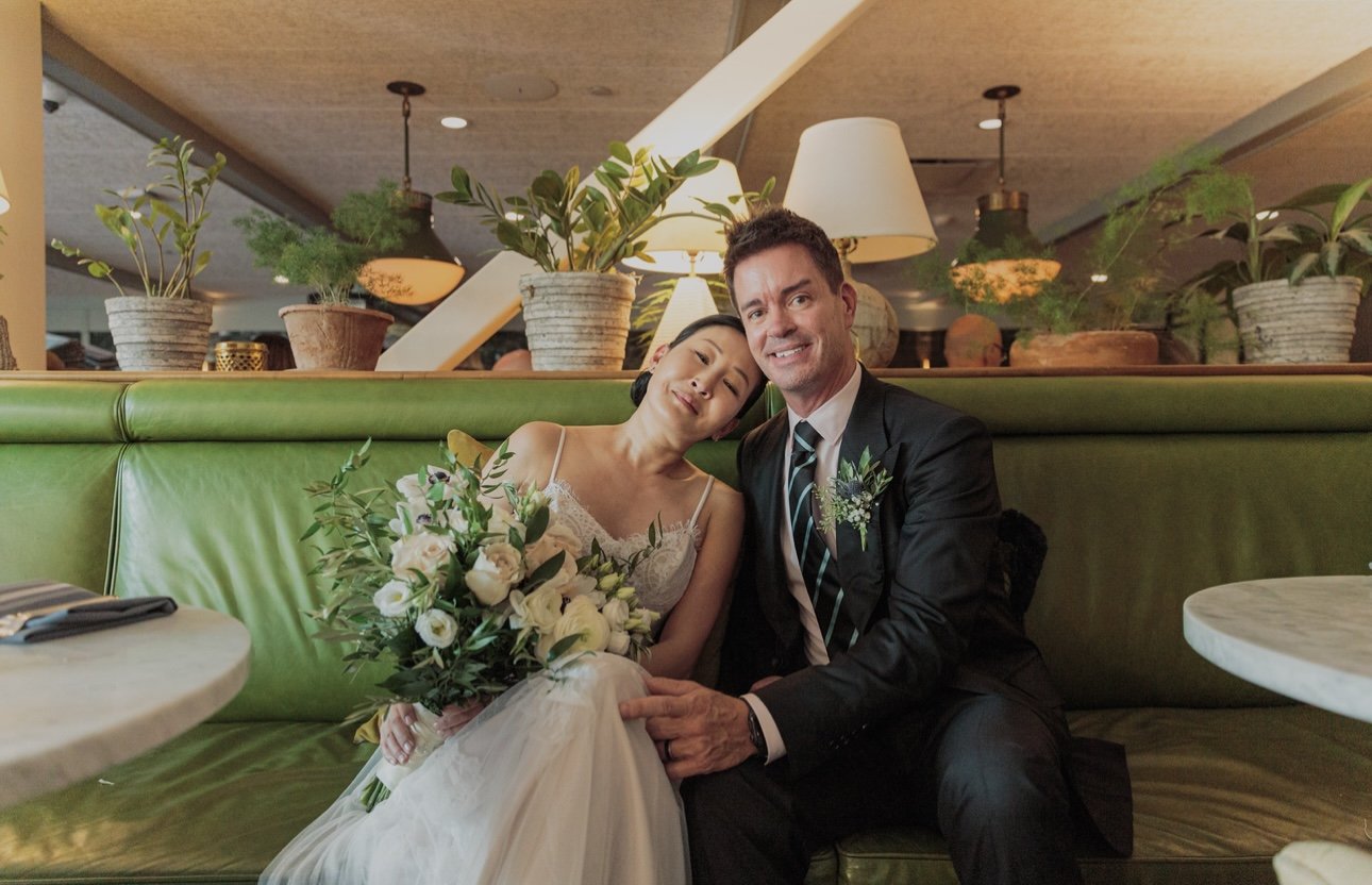 A bride and groom sitting on a green booth in a restaurant or cafe, posing happily for a photo. The bride is holding a bouquet of flowers and wearing a white wedding dress, while the groom is dressed in a black suit with a striped tie and a boutonniere. They are smiling and leaning close to each other amidst warm lighting and potted plants in the background.