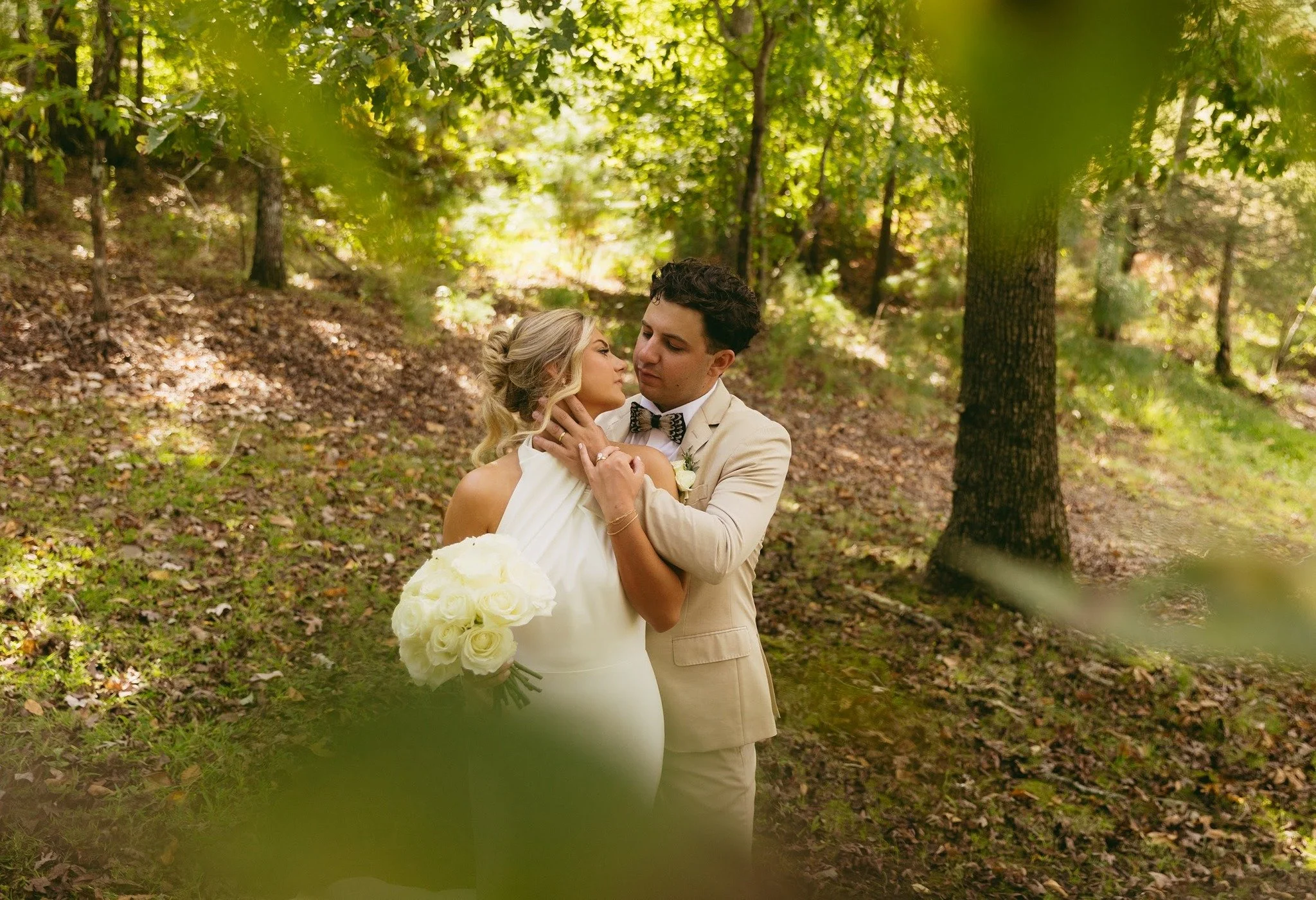 A bride and groom sharing a loving moment in a forested area during their wedding, with the bride holding a bouquet of white roses.