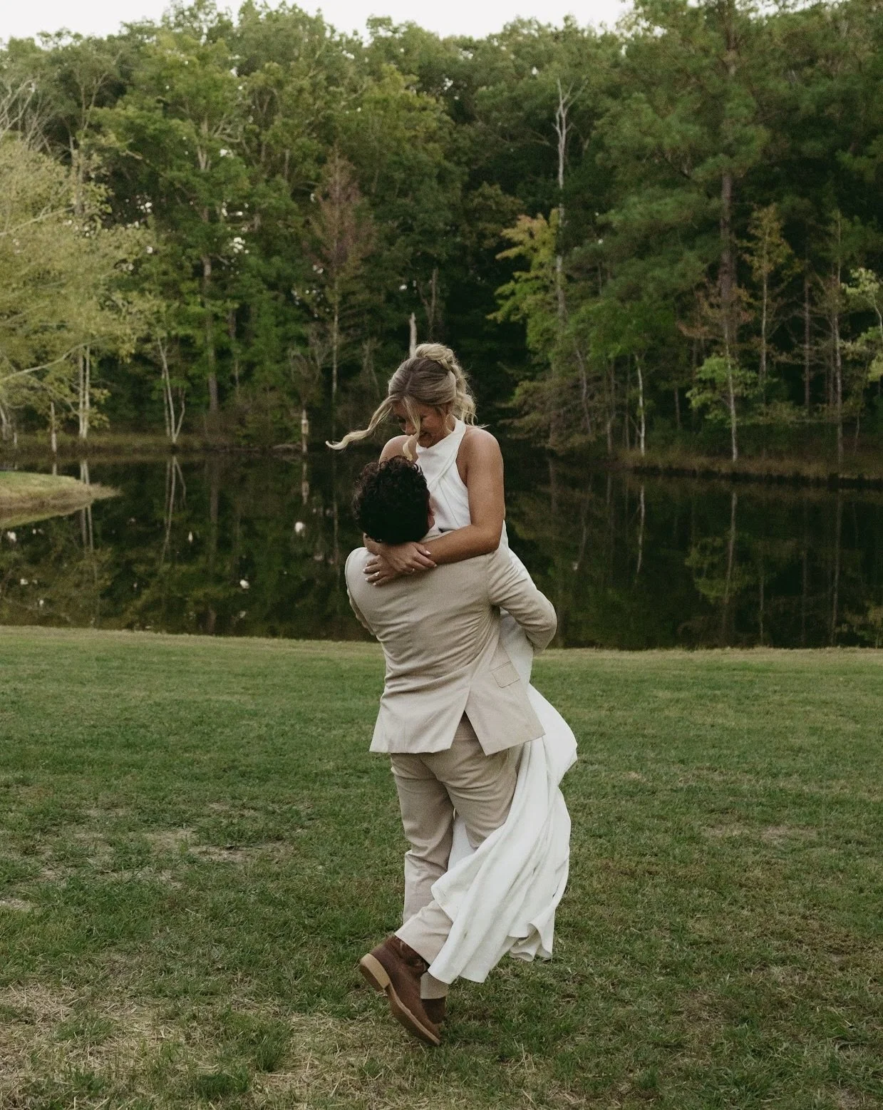 A couple is outdoors near a lake, with the man lifting the woman in his arms. The woman is wearing a white dress, and the man is dressed in a beige suit. The background features a forest with green trees and the lake reflecting the surroundings.