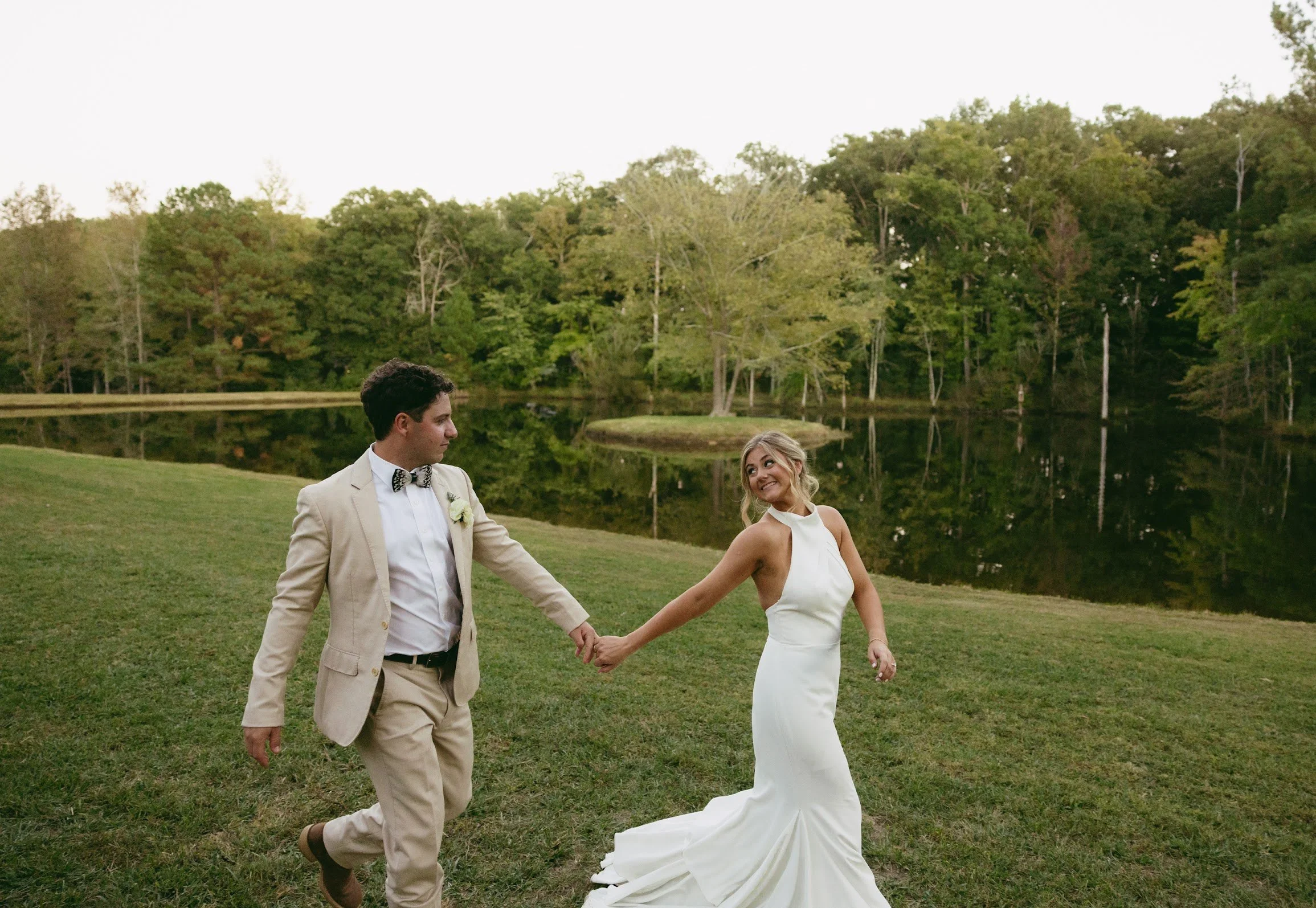 A bride and groom holding hands and dancing outdoors near a lake with trees in the background.