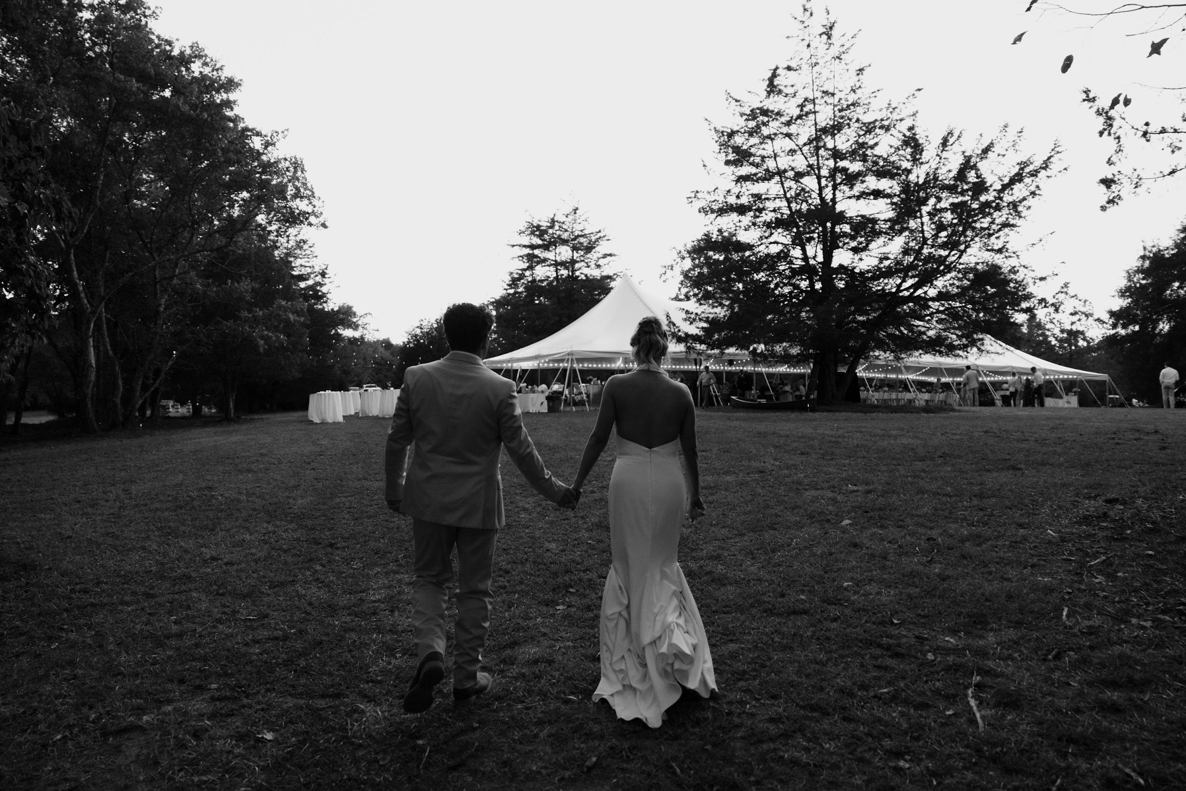 A couple dressed in wedding attire walking hand in hand towards a large outdoor tent at sunset, with trees surrounding the area.