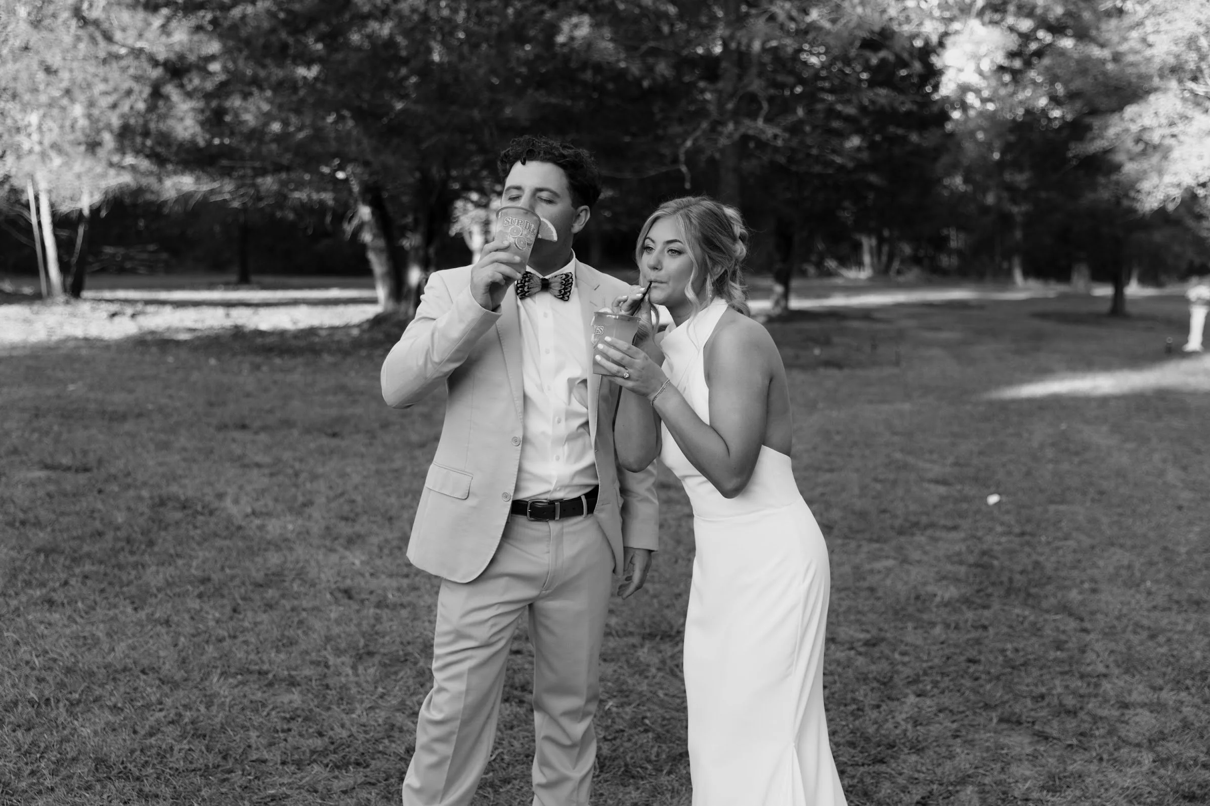A black and white photo of a man and woman in wedding attire in a park, both sipping drinks with straws. The man wears a suit and bow tie, and the woman wears a sleeveless wedding dress.