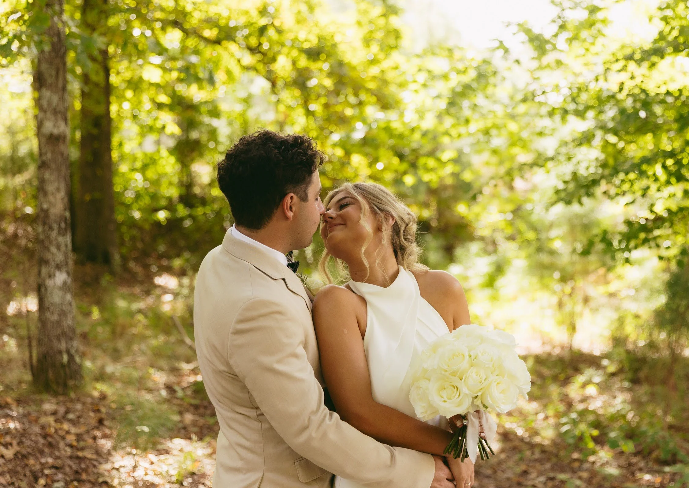 A bride and groom embrace in a forest with sunlight filtering through green trees, the bride holding a bouquet of white roses.