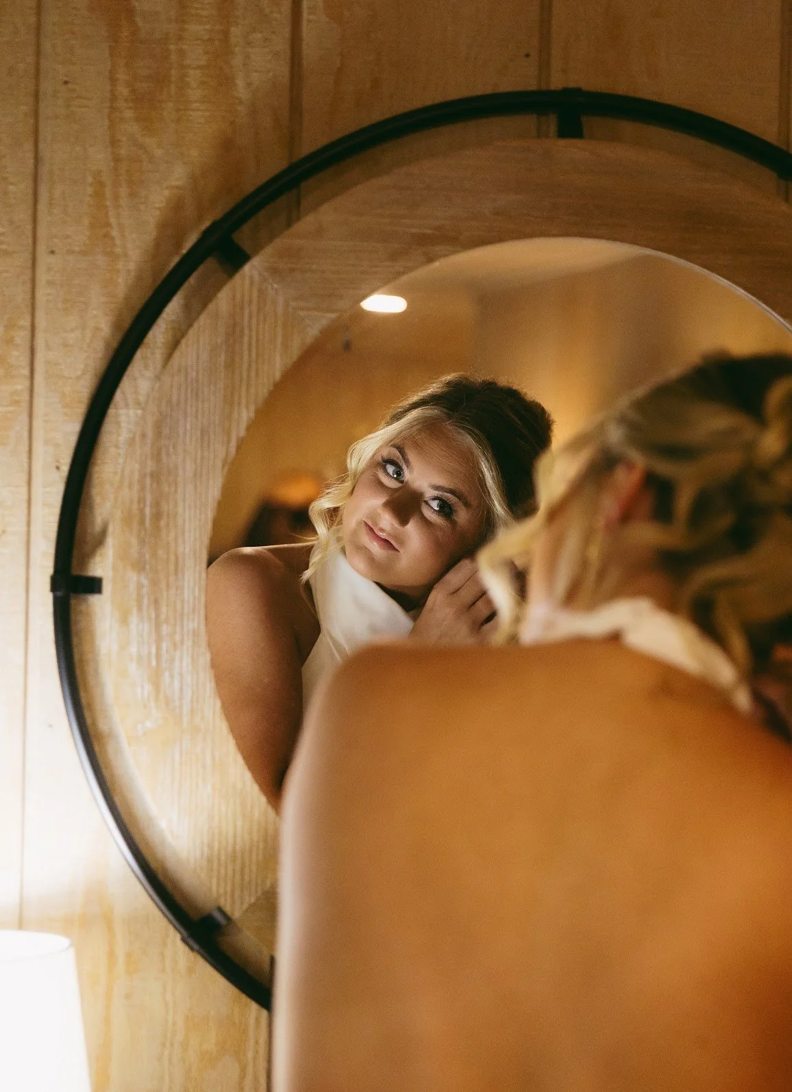A woman looks at her reflection in a circular mirror mounted on a wooden wall, with warm ambient lighting illuminating the scene.