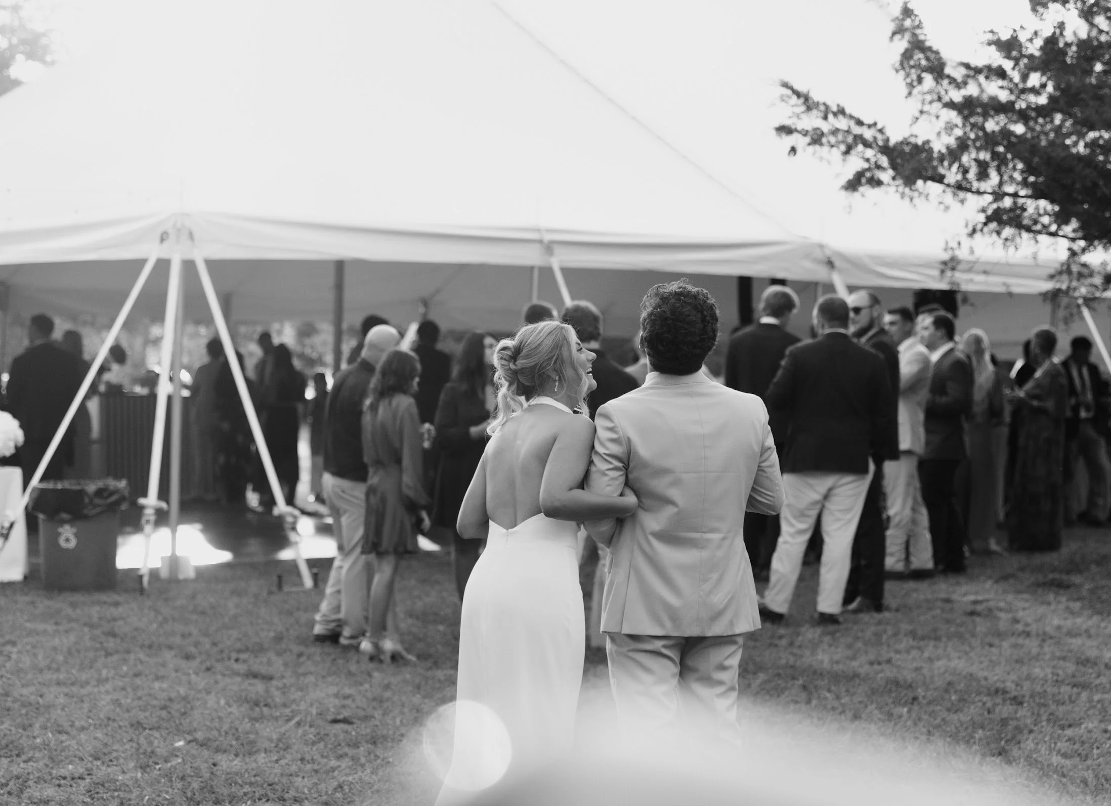 A bride and groom walking towards a gathering of guests at an outdoor wedding reception under a large white tent.