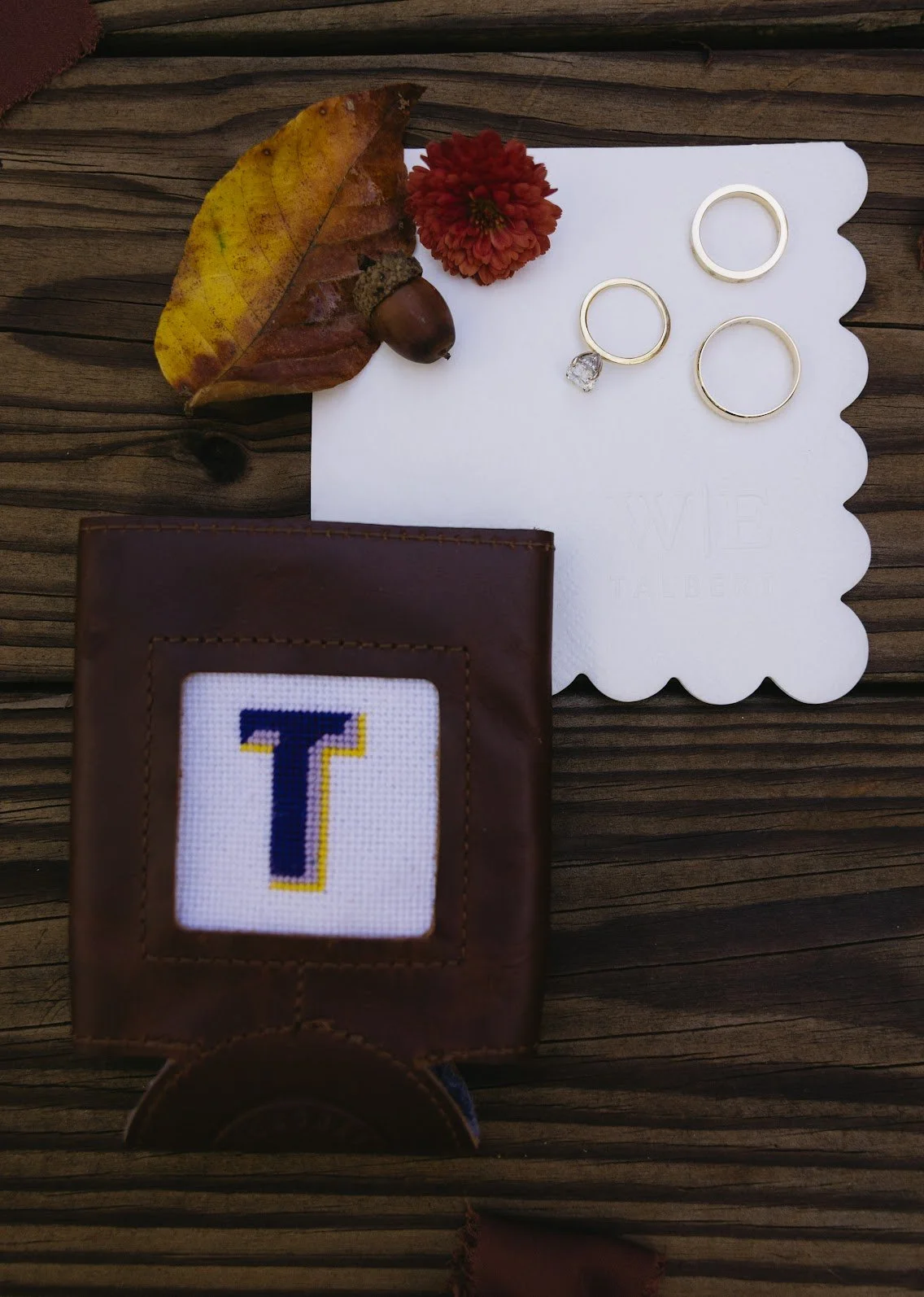 A leather wallet with a cross-stitch letter T, a white card with jewelry, a yellow and brown leaf, a red flower, and an acorn on a wooden surface.