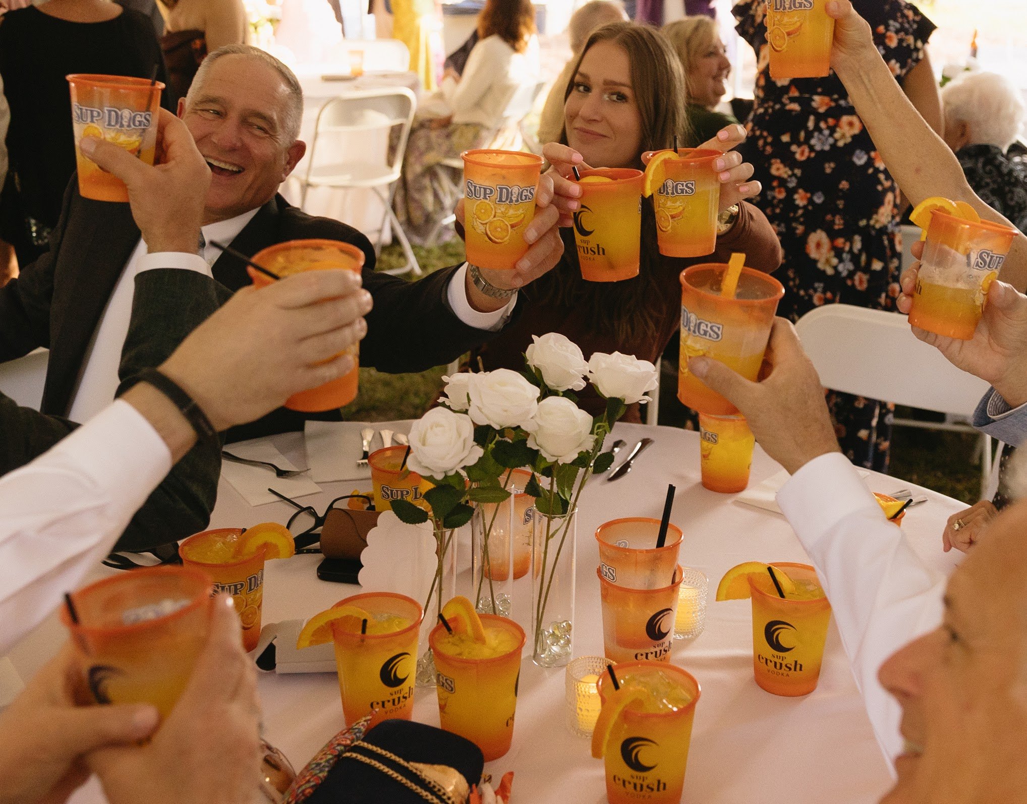People holding up cups with orange and yellow labels, sitting around a table with white roses and candles, celebrating at a party.
