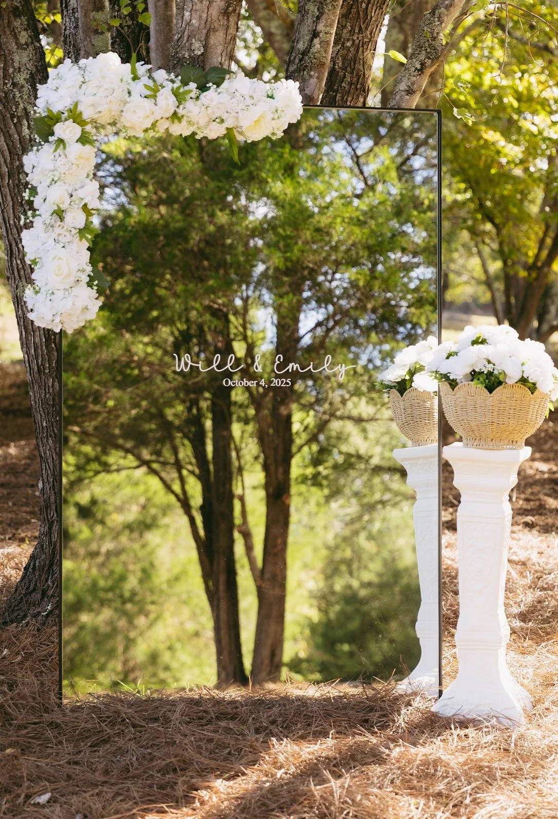 Outdoor wedding mirror with white floral decorations and two white pedestals holding baskets of white flowers, trees and greenery in the background.