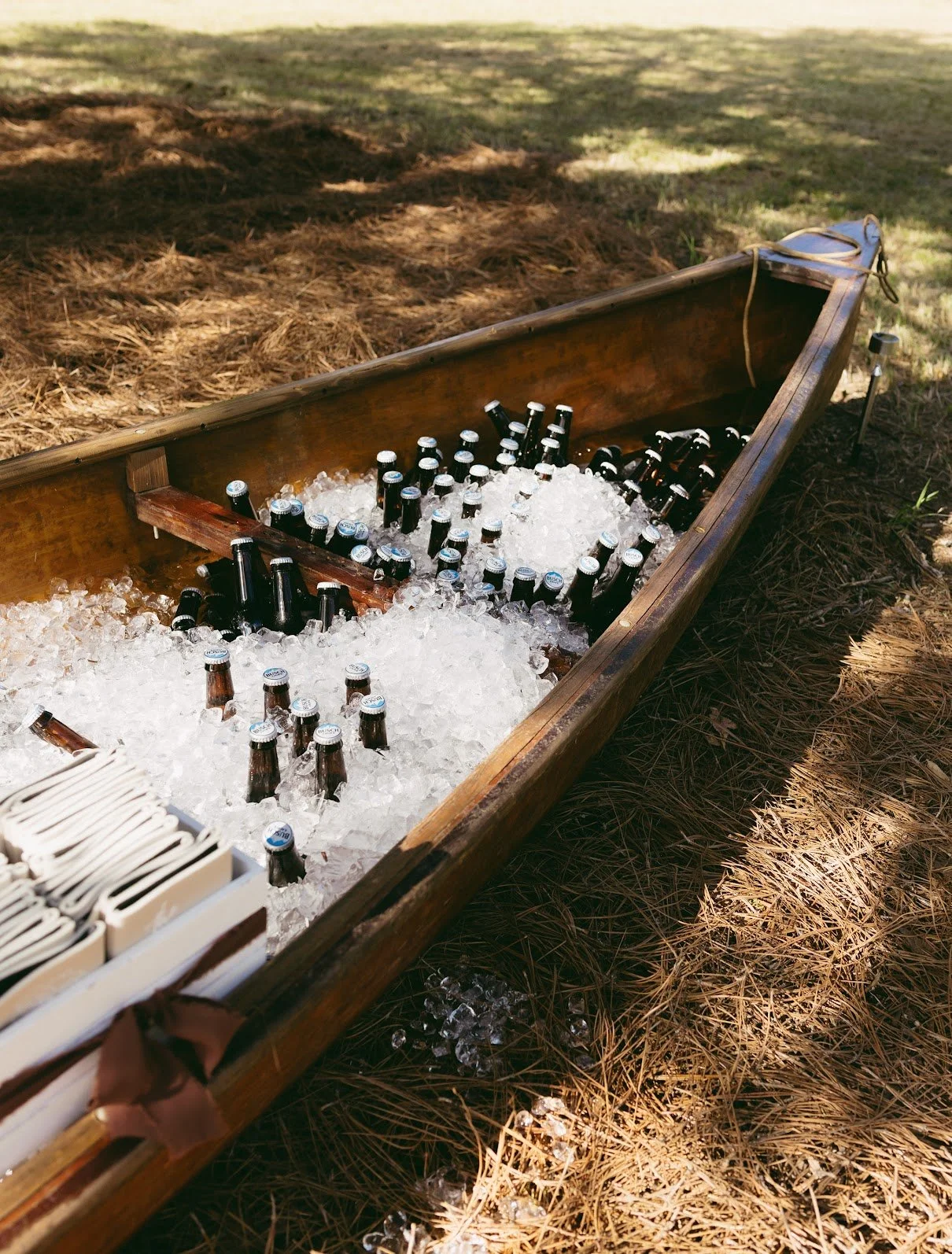 A wooden canoe filled with ice and bottles of beer, resting on a grassy area with pine needles on the ground.