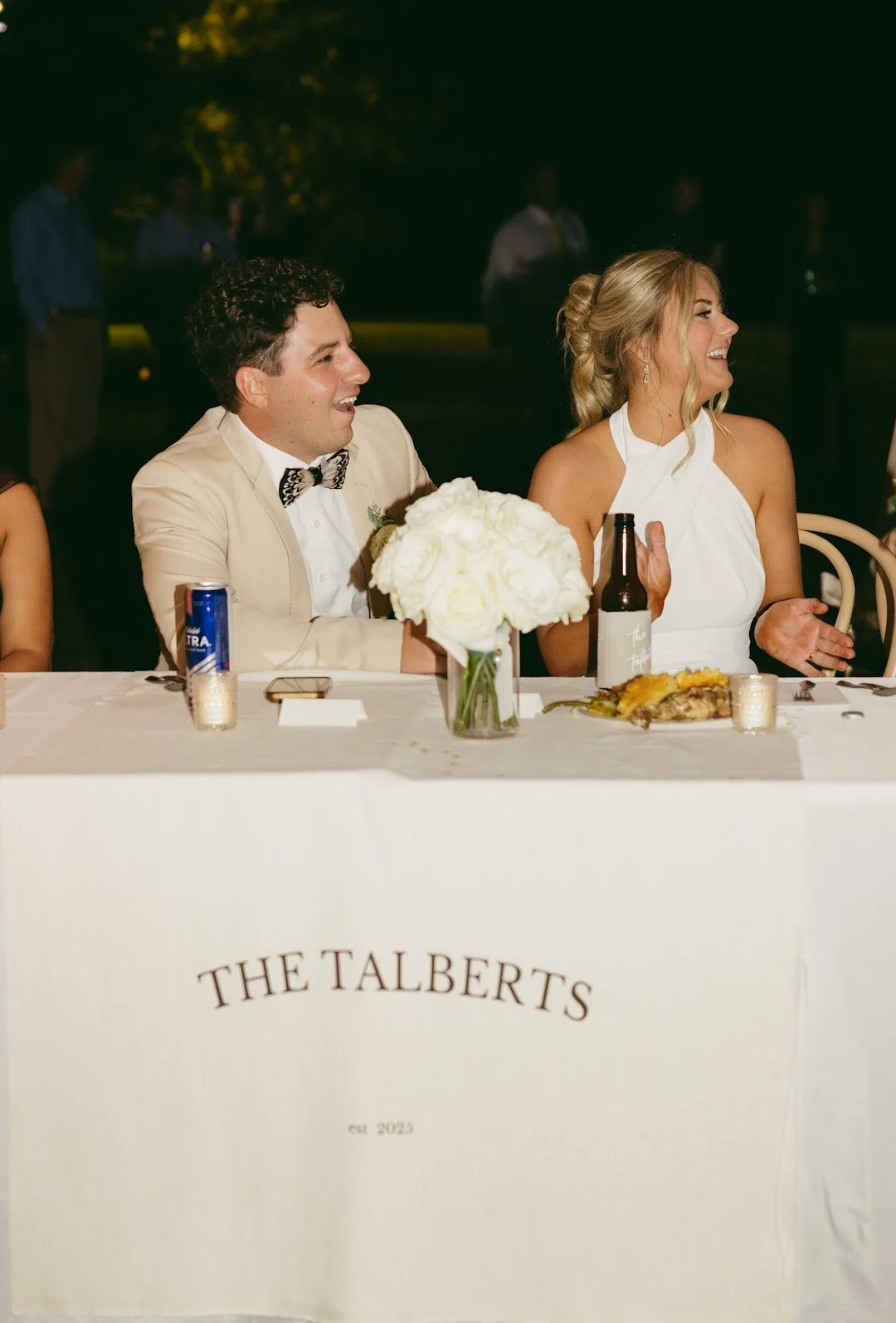 At a wedding reception, a man in a beige tuxedo with a bow tie and a woman in a white dress are sitting at a decorated table, laughing. The table has a white tablecloth, a floral centerpiece, and drinks including a can and a bottle. The tablecloth ha