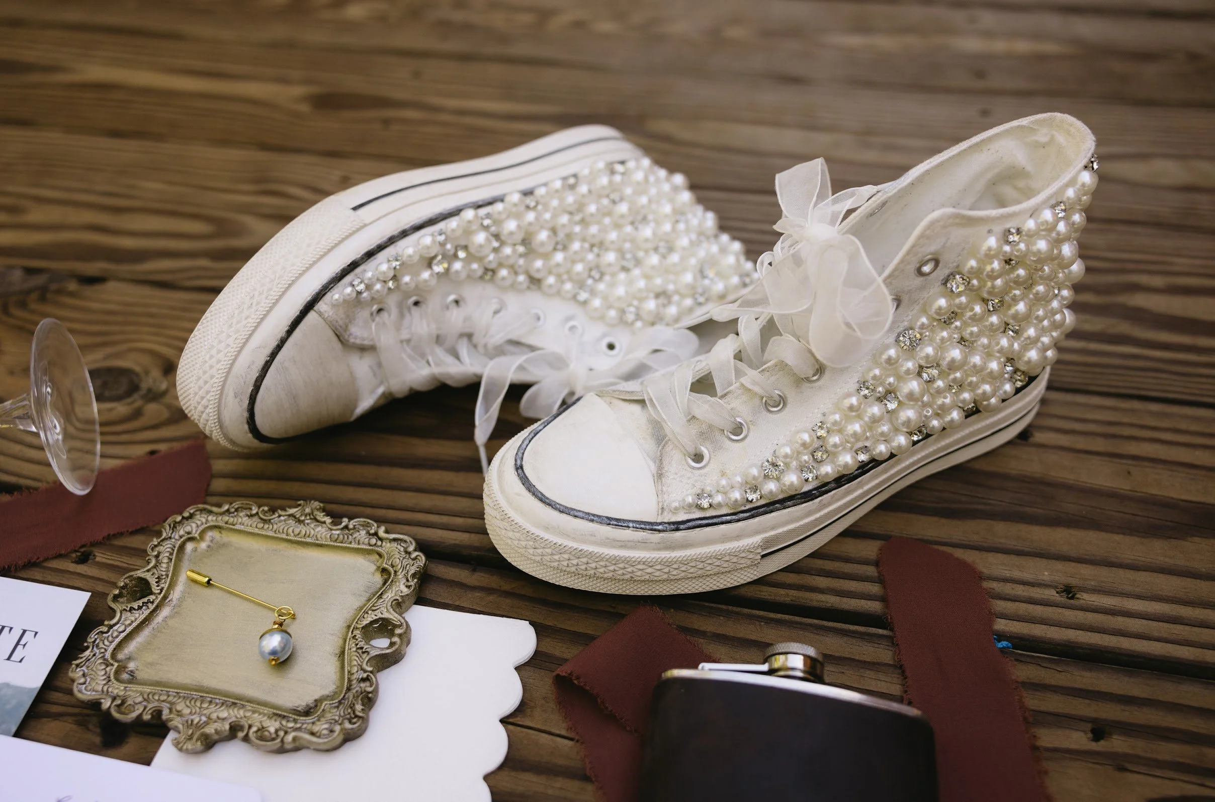 White sneakers decorated with pearls and rhinestones, placed on a wooden surface surrounded by jewelry and accessories.