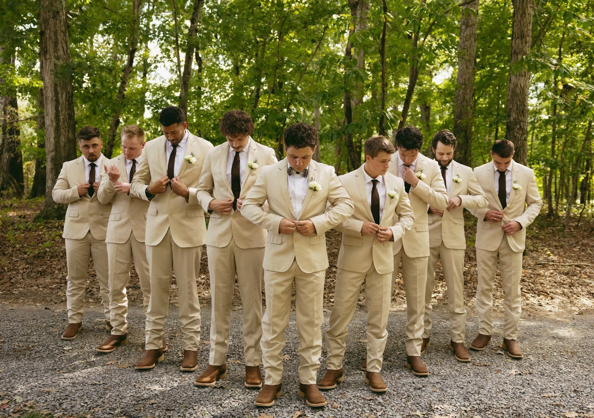 A group of nine men dressed in beige suits, white shirts, brown shoes, and ties, standing outdoors in a wooded area adjusting their suit jackets.