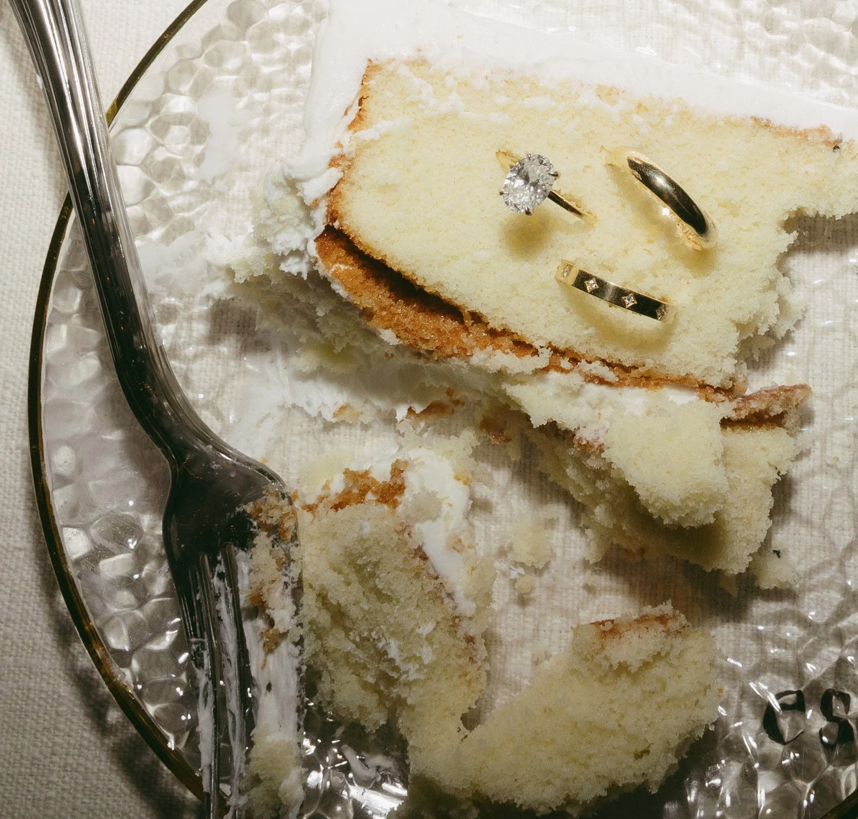 Pieces of a broken white cake on a decorative glass plate with three rings placed on top, and a metal fork resting on the side.