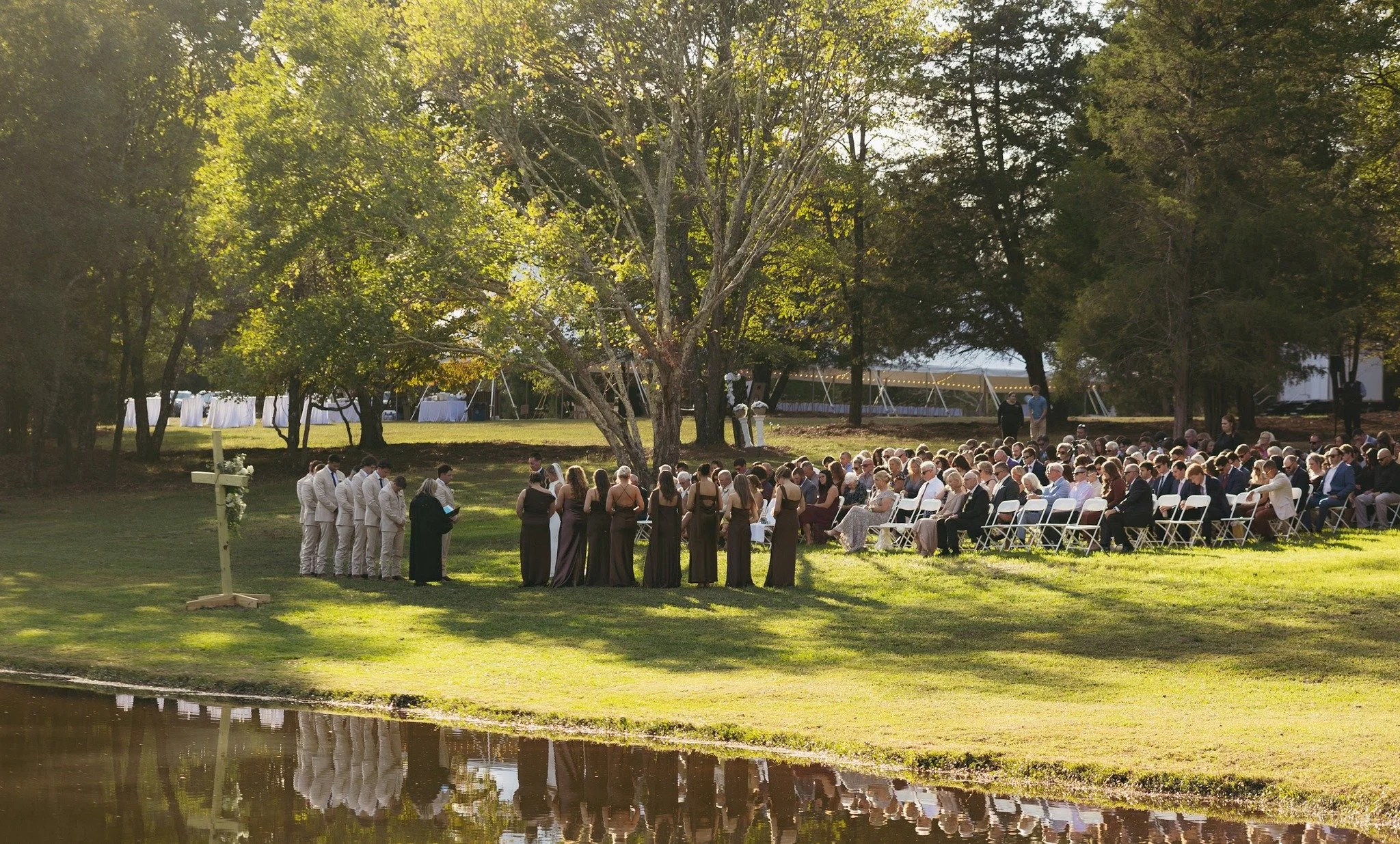 Outdoor wedding ceremony with guests seated on white chairs, wedding party standing at the altar, officiant reading, in a park-like setting with trees and reflections in a pond.