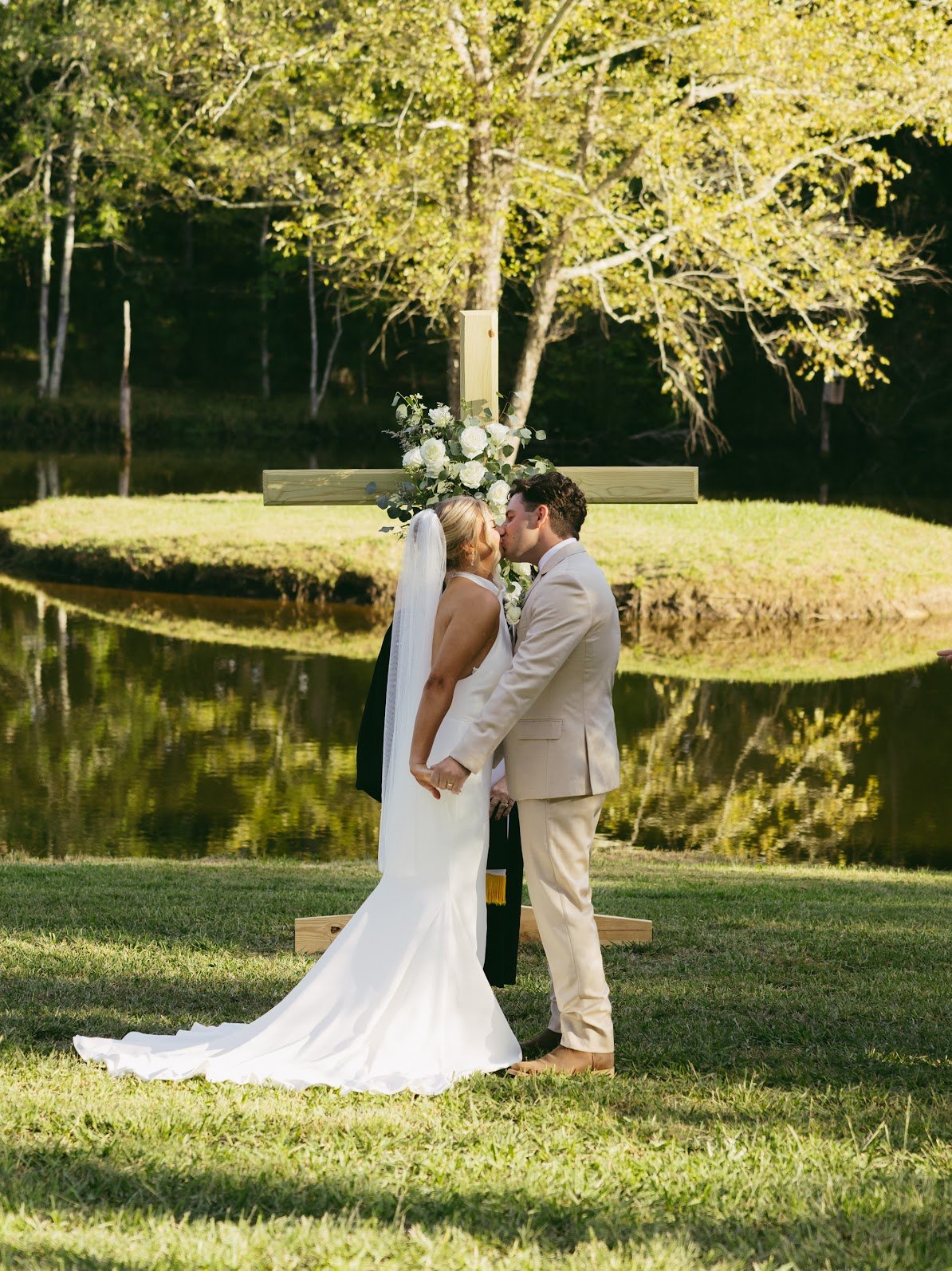 A bride and groom kiss in front of a wooden cross decorated with white flowers and greenery, near a pond with trees in the background on a sunny day.