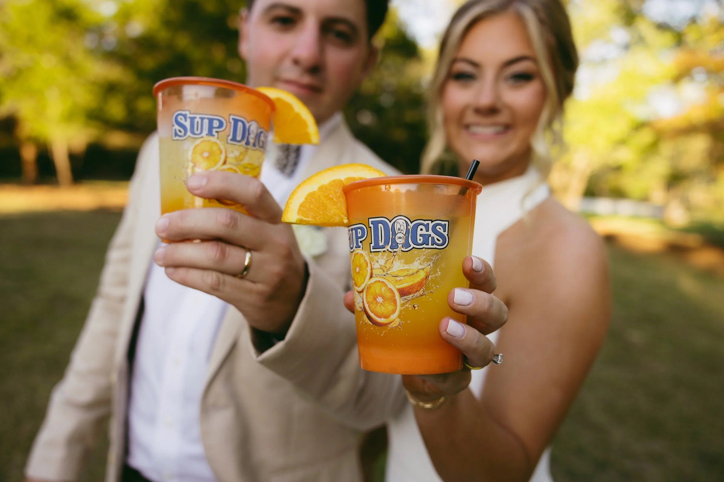 A couple holding colorful cups with orange slices outdoors in a park on a sunny day.