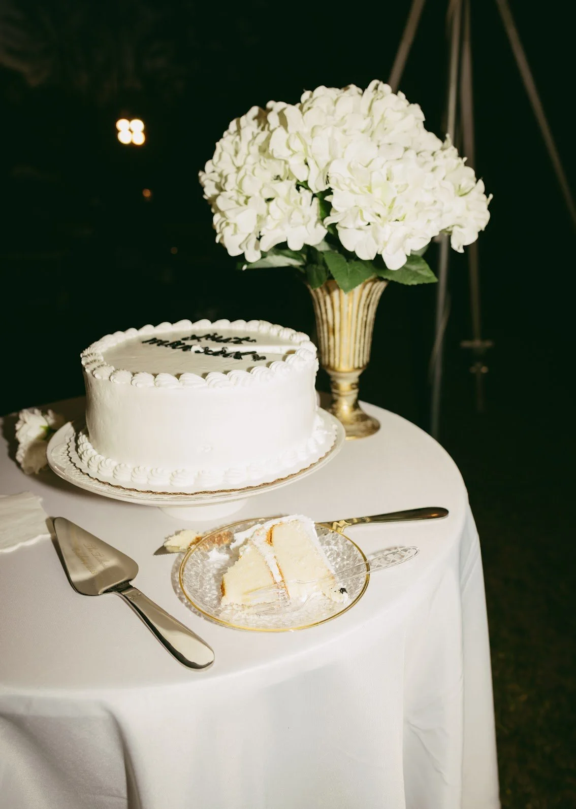 A wedding cake with white frosting and decorative borders, placed on a white tablecloth. There is a partially eaten slice of cake on a clear plate with a gold rim, and cake serving utensils on the table. A large white hydrangea flower arrangement in 