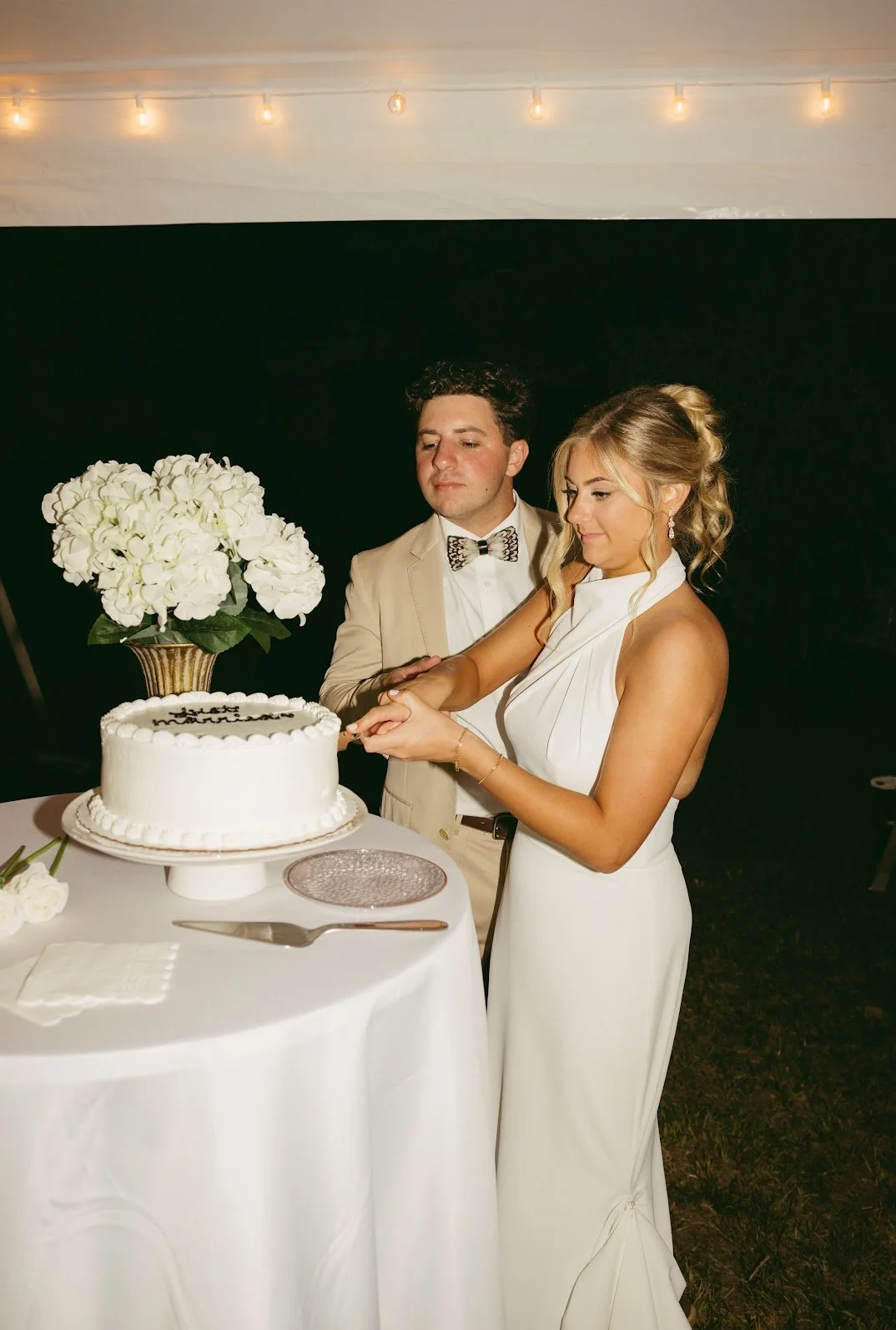A newlywed couple cutting a wedding cake together at their reception, with the bride wearing a white dress and the groom in a tan suit and bow tie, on a table with a large bouquet of white flowers.