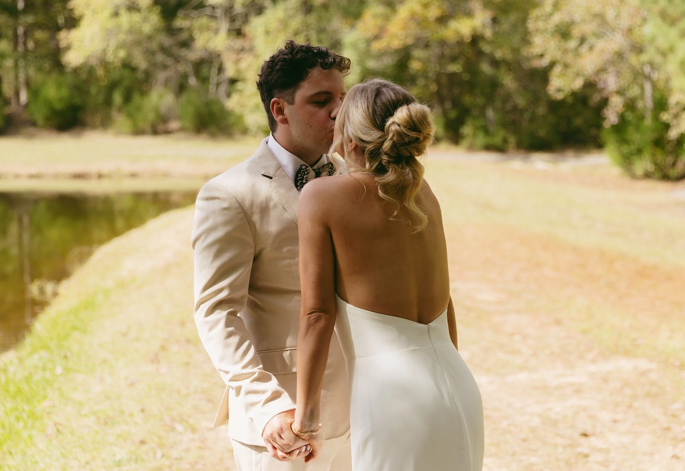 A couple in wedding attire holding hands and sharing a kiss outdoors near a body of water, with trees and greenery in the background.