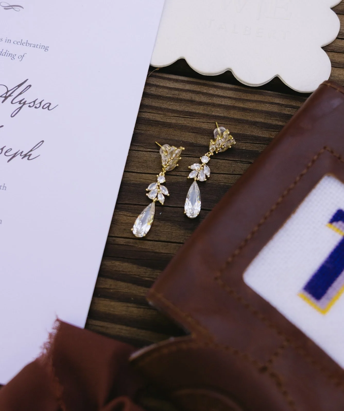 Wedding invitation, pearl and crystal earrings, brown leather wallet, and wooden surface.