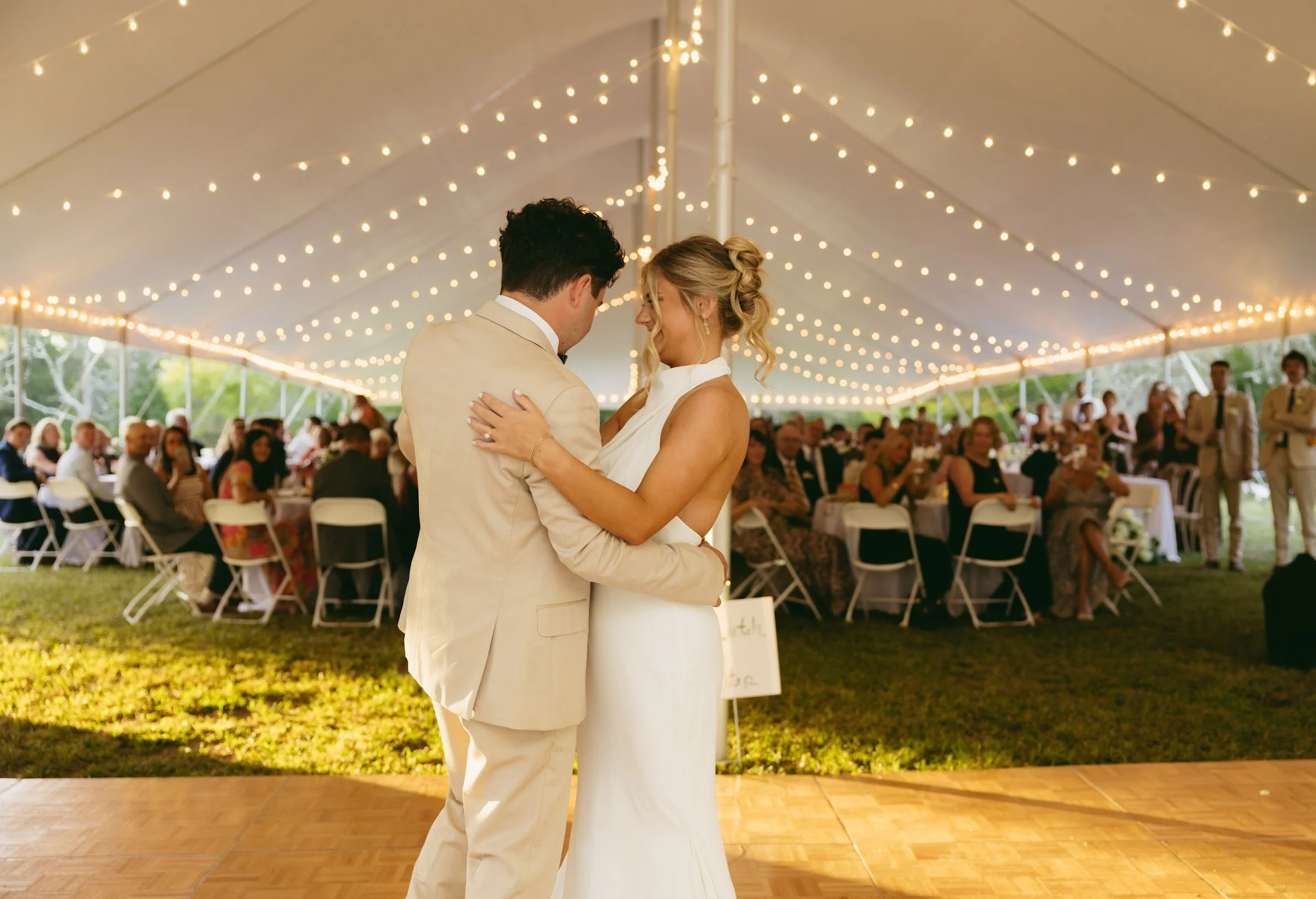 A bride and groom sharing their first dance under a large tent with string lights, surrounded by seated wedding guests.