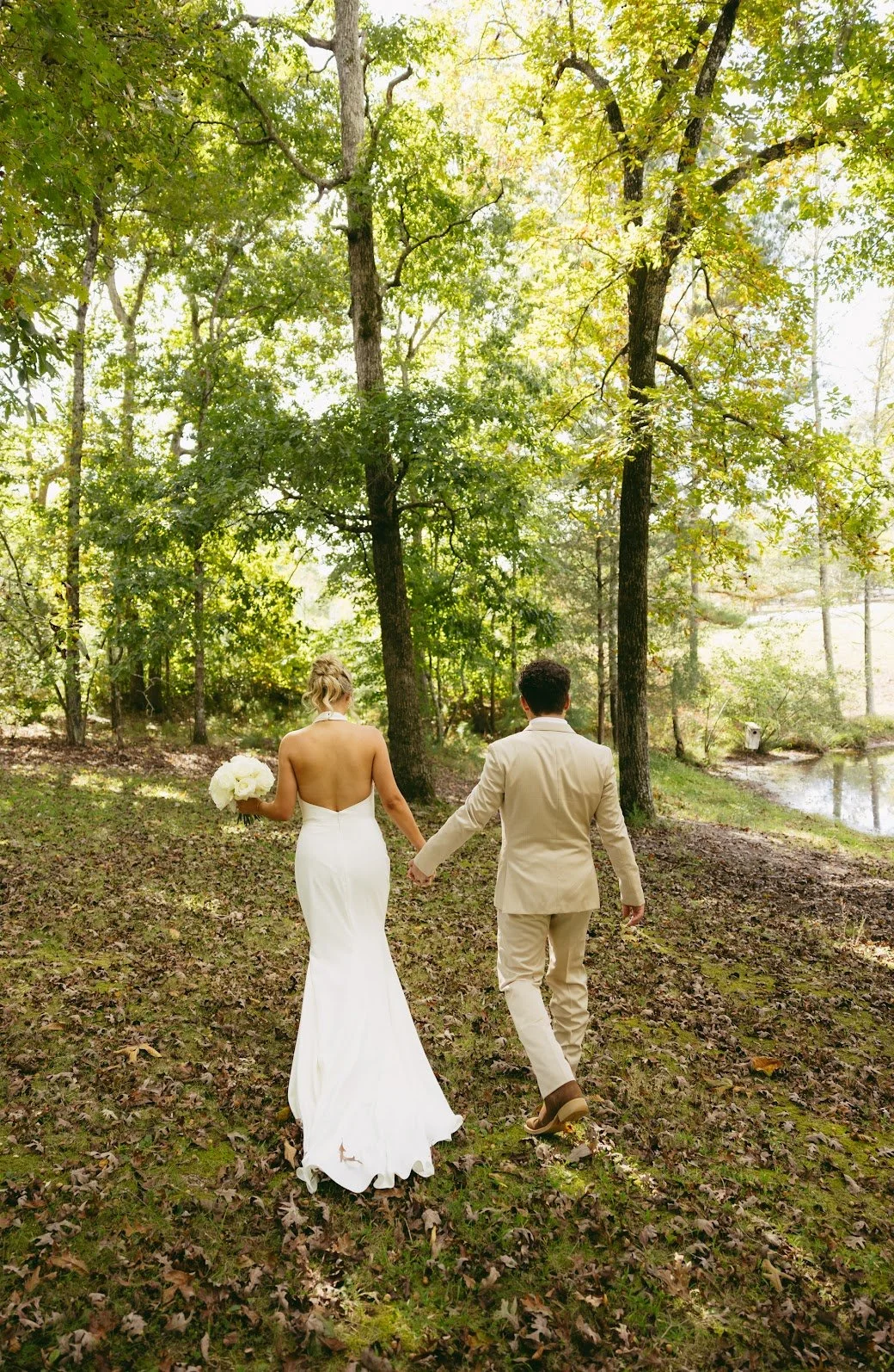 A bride and groom walk hand in hand through a wooded area with green trees and sunlight filtering through the branches. The bride wears a white, strapless wedding gown and holds a bouquet of white flowers, while the groom wears a beige suit.