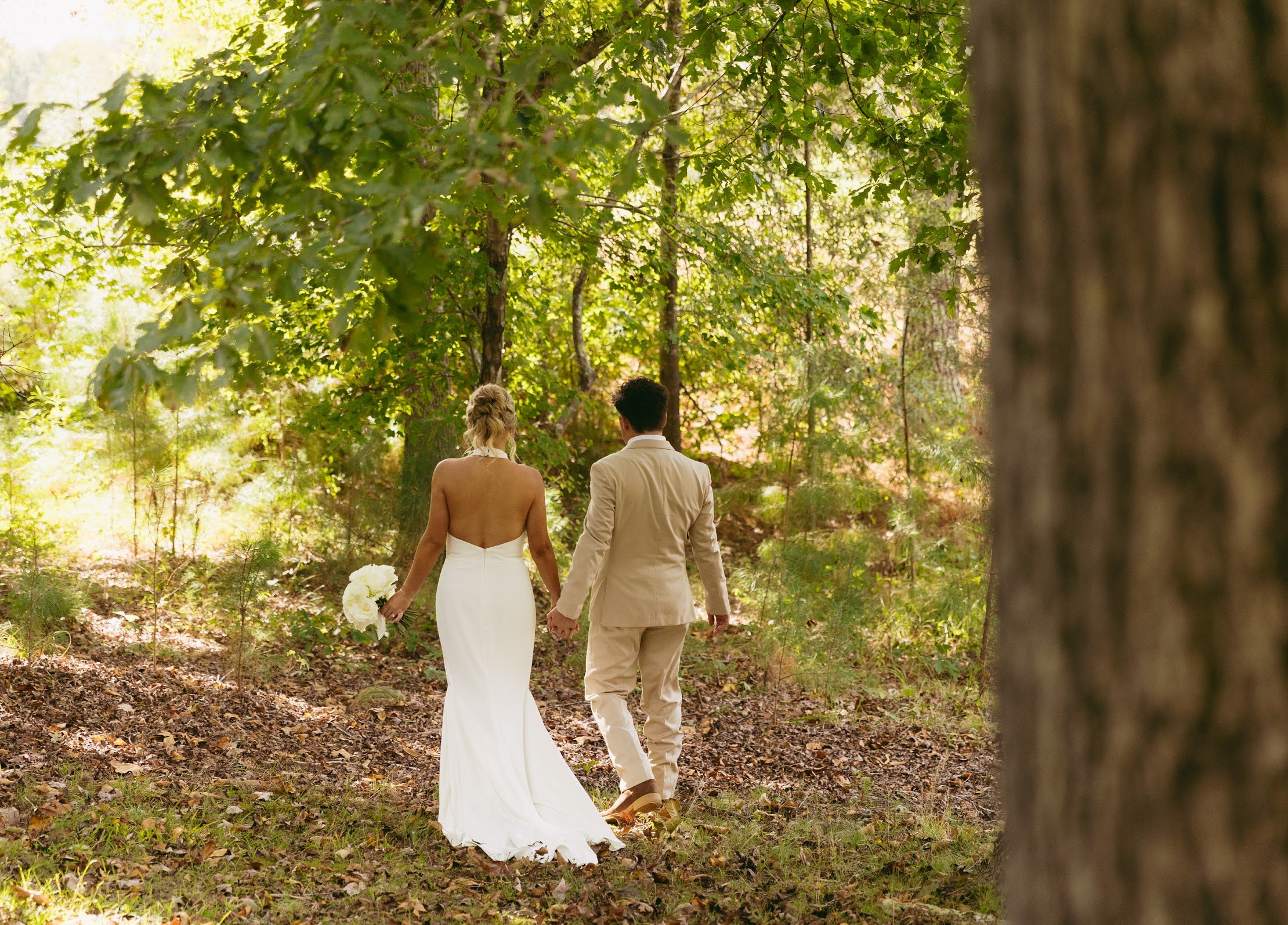 A bride and groom walking hand in hand through a wooded forest on their wedding day, with the bride holding a bouquet of white flowers.