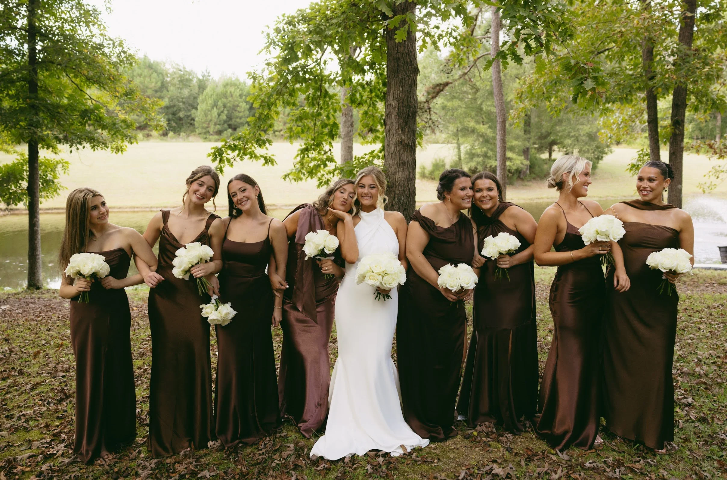 A bride in a white dress with blonde hair standing with eight bridesmaids in dark brown dresses holding bouquets of white flowers outdoors near a pond and trees.