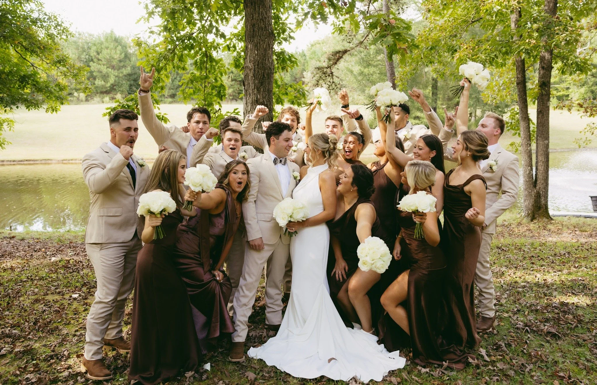 Wedding celebration outdoors with group of people, bride and groom at center, surrounded by bridesmaids and groomsmen, holding white bouquets, all smiling and cheering, in a park with trees and water in the background.