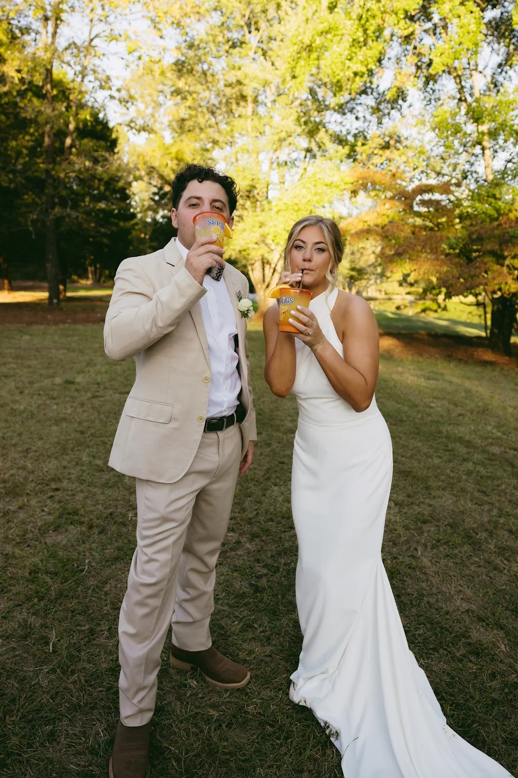 A man and a woman in wedding attire standing outdoors in a park, holding cups labeled 'SUP' and 'DIPS'. They are drinking from straws, with trees and sunlight in the background.