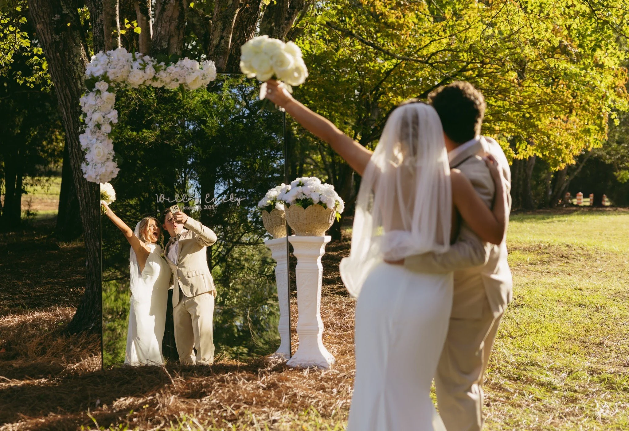 A bride and groom embrace outdoors during their wedding, with a large mirror reflecting the bride holding a bouquet and a woman taking a photo, surrounded by green trees and floral decorations.