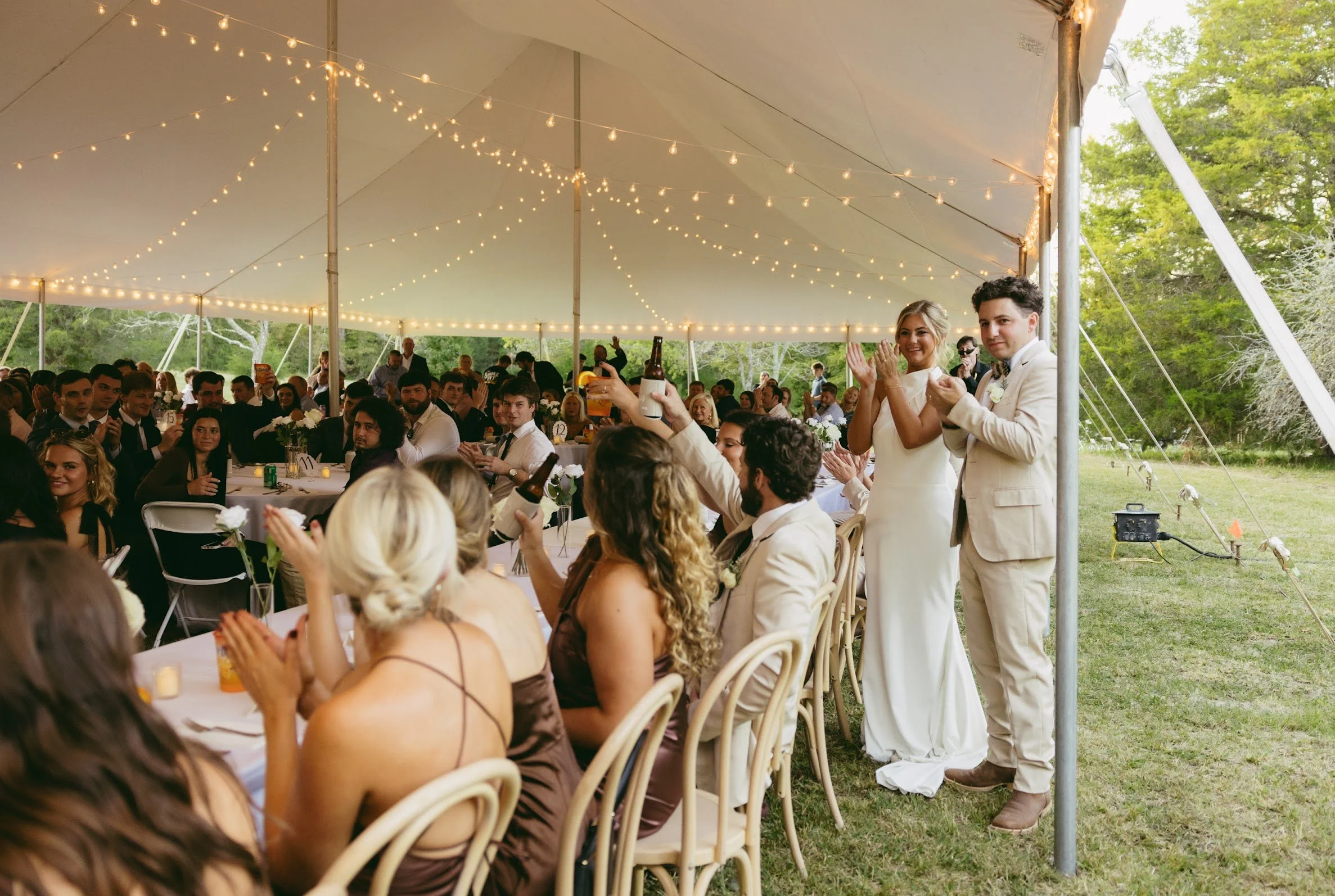 A wedding reception under a large white tent with string lights, featuring a bride and groom standing together, holding drinks, with guests seated at long tables outside.