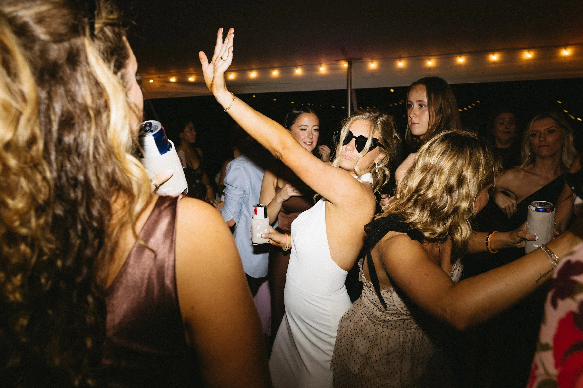 Women dancing and socializing at a party or event, with some holding cans and cups, under string lights.