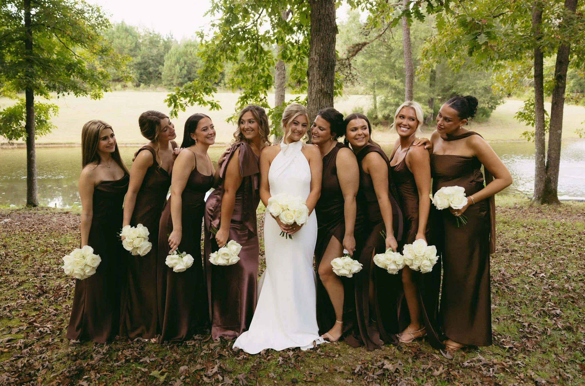A bride and her eight bridesmaids standing outdoors by a lake and trees, all smiling and holding white bouquets. The bride wears a white gown, the bridesmaids wear brown dresses.