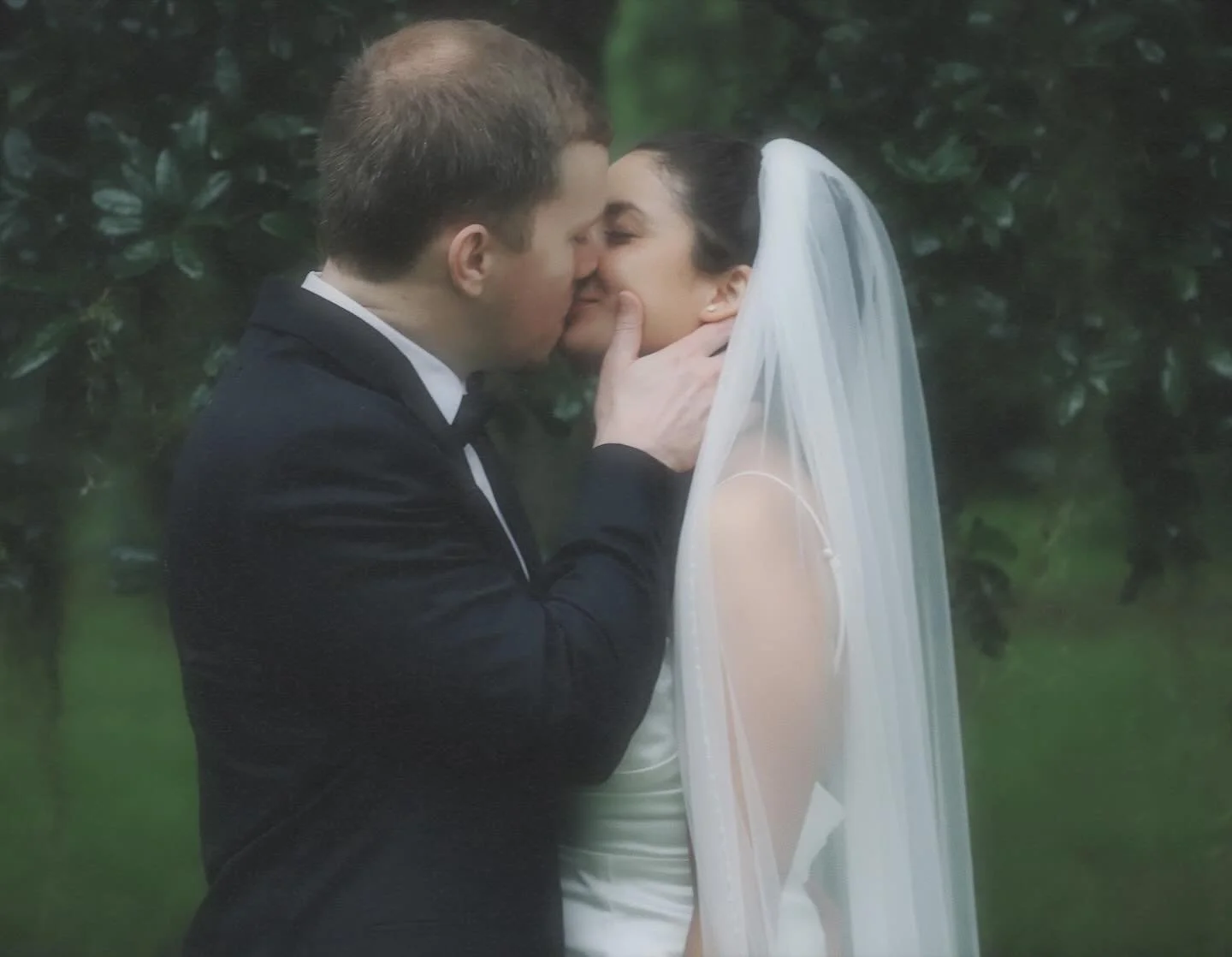 A couple dressed in wedding attire sharing a kiss outdoors, surrounded by greenery.
