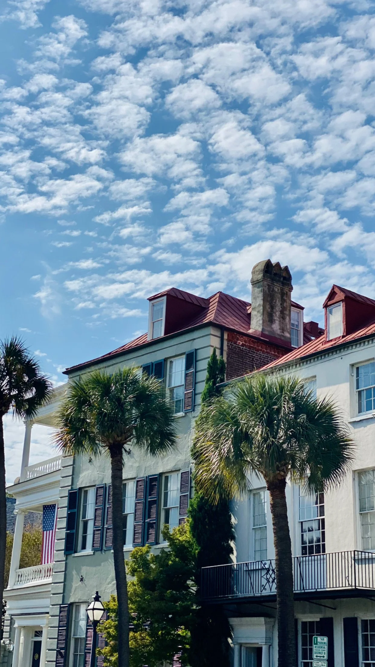 A residential street with multi-story houses featuring large windows and colorful shutters, under a partly cloudy blue sky, with palm trees and a streetlamp in the foreground.