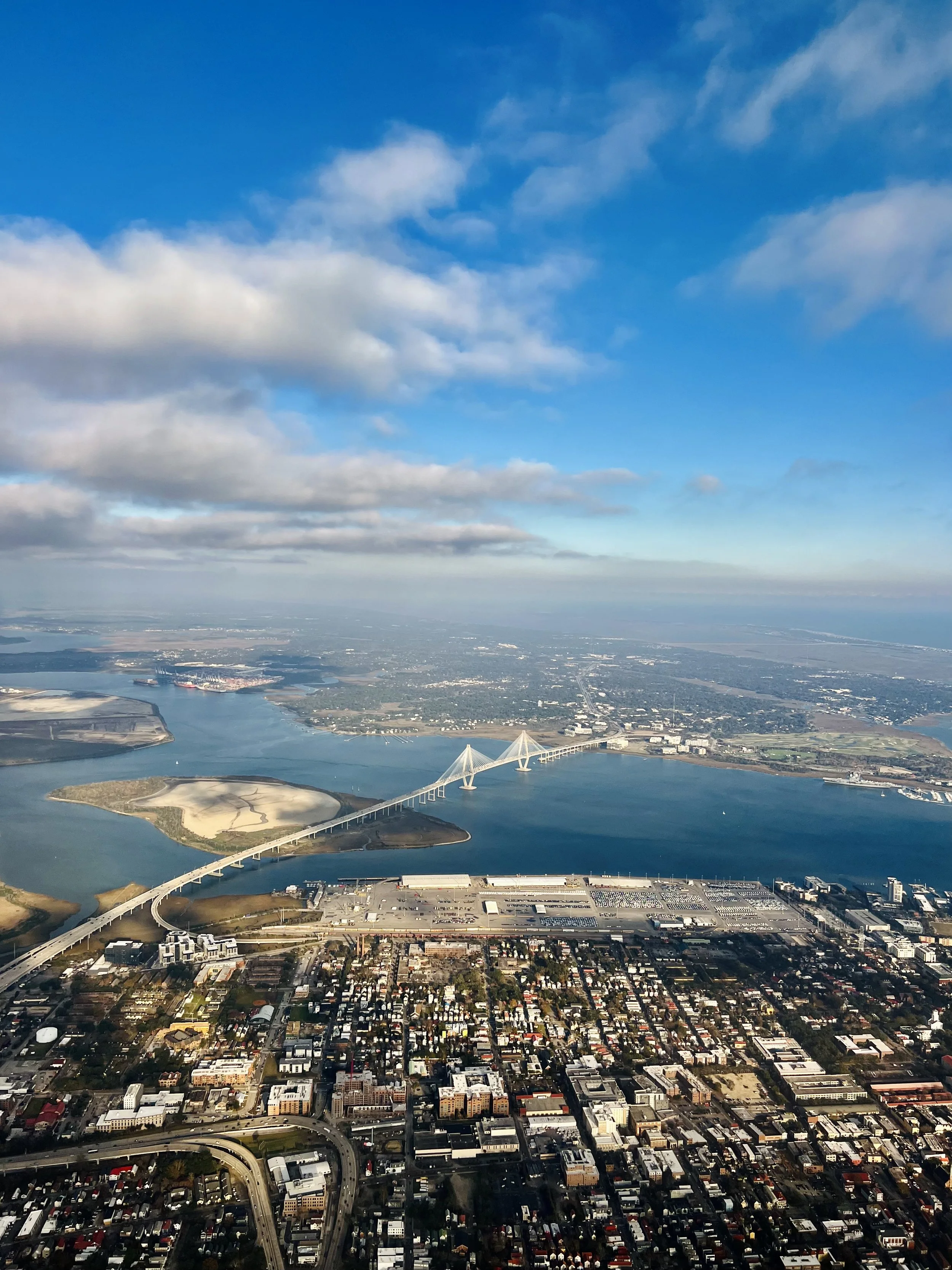 Aerial view of a city with a bridge crossing a body of water, with a mix of urban and natural areas under a partly cloudy sky.