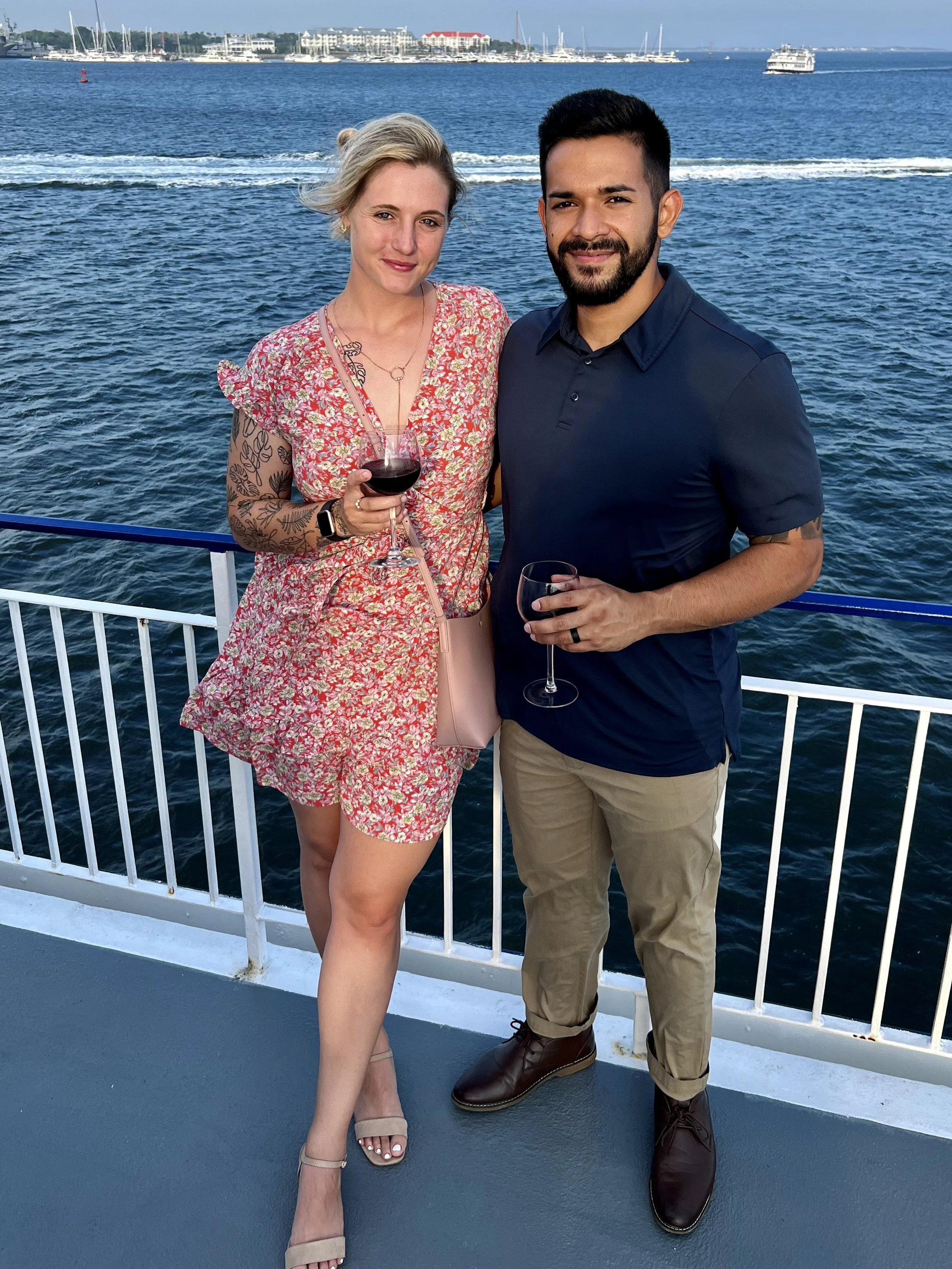 A woman and a man standing on a boat deck by the water, holding glasses of red wine, with boats and a city skyline in the background.