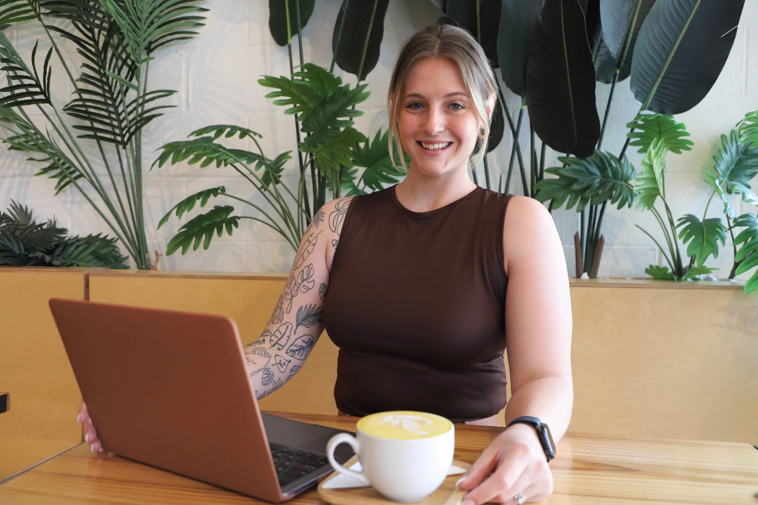 A smiling woman with blonde hair, wearing a sleeveless brown top and a tattoo on her left arm, sitting at a wooden table with a pink laptop and a cup of coffee, surrounded by green plants.