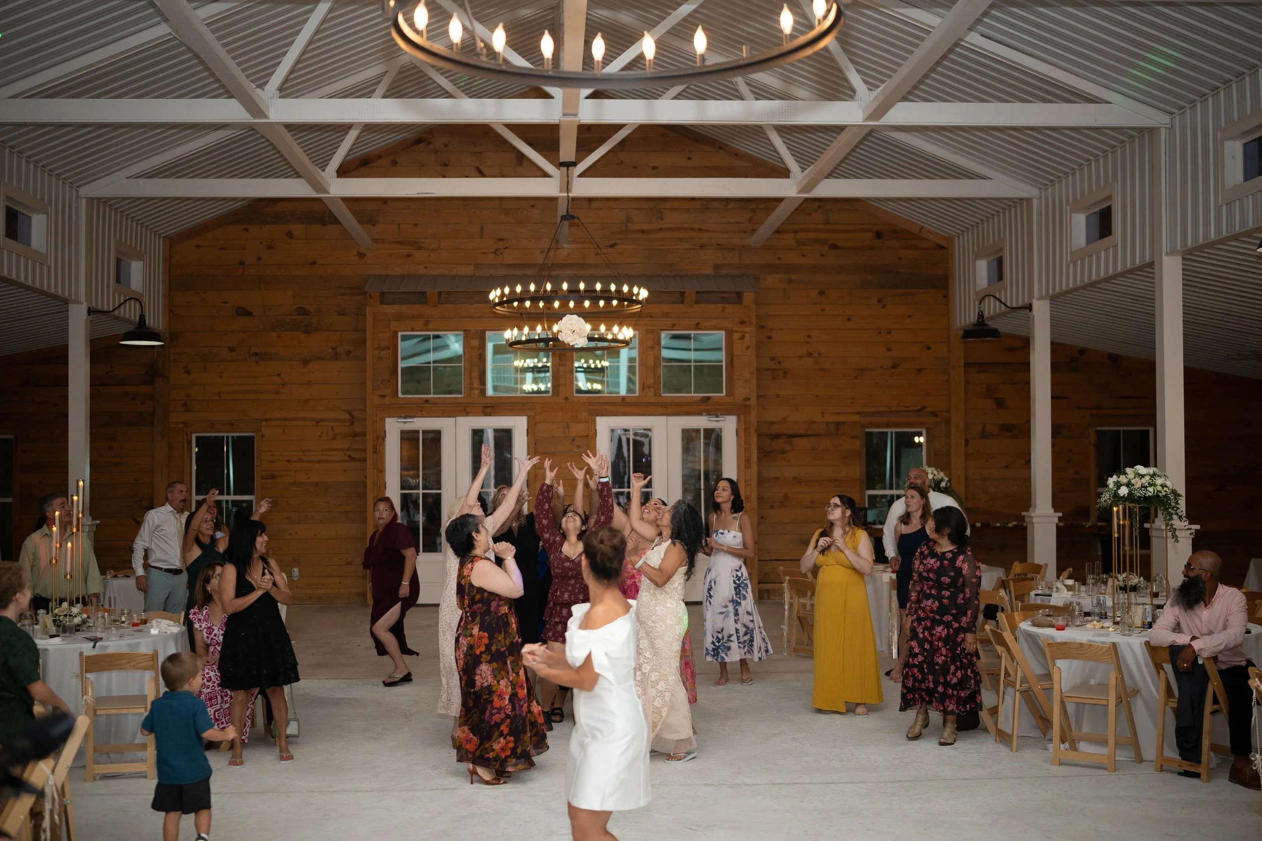 Guests dancing and socializing at a wedding reception inside a barn-style venue with wooden walls and high ceiling, decorated with chandeliers and floral arrangements.